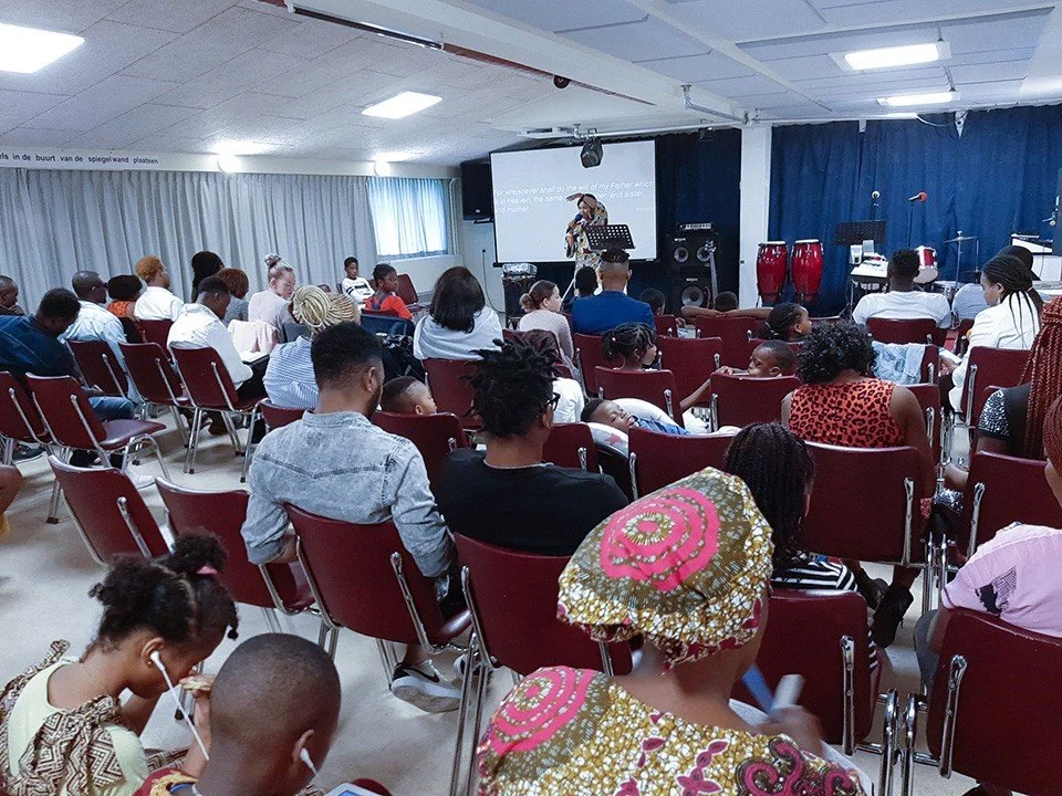Group of people attending a church service or event, sitting on red chairs, facing a speaker at the front in a room with blue curtains and a large screen, colorful drums and musical instruments visible.