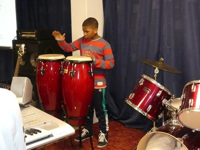 A young boy playing drums in a room with musical instruments, including congas and a drum kit, with a keyboard in the foreground.