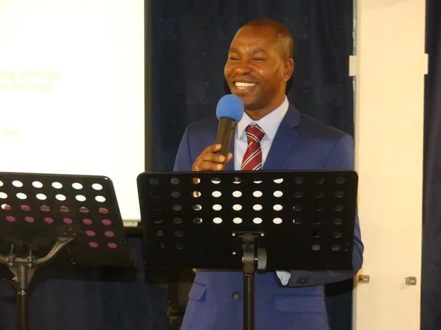 A man in a suit smiling and speaking into a blue microphone during a presentation or speech.