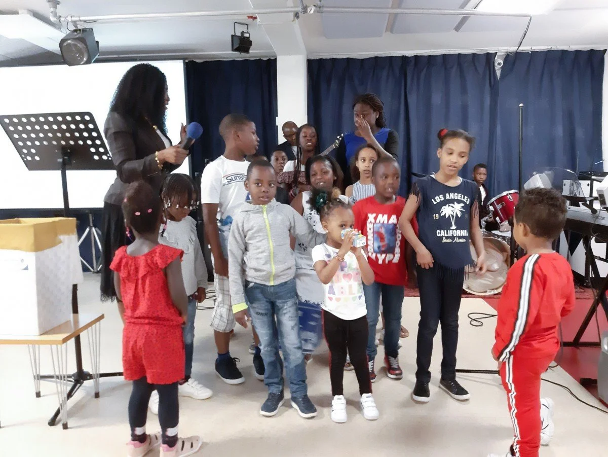A group of children standing on a stage, some singing or talking, with two women, one holding a microphone, in a room with music instruments and a dark blue curtain in the background.