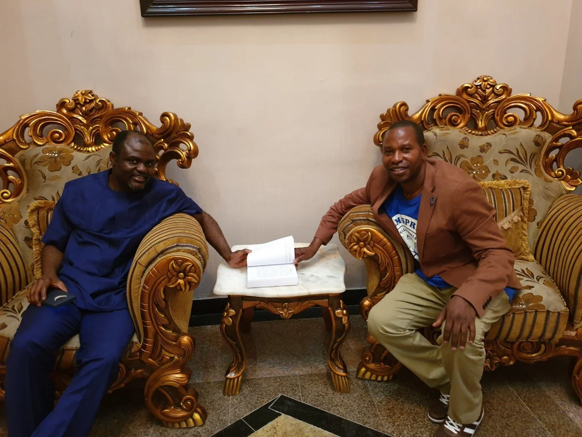Two men sitting on ornate gold and patterned armchairs, holding open books placed on a small white table between them in a room with beige walls.