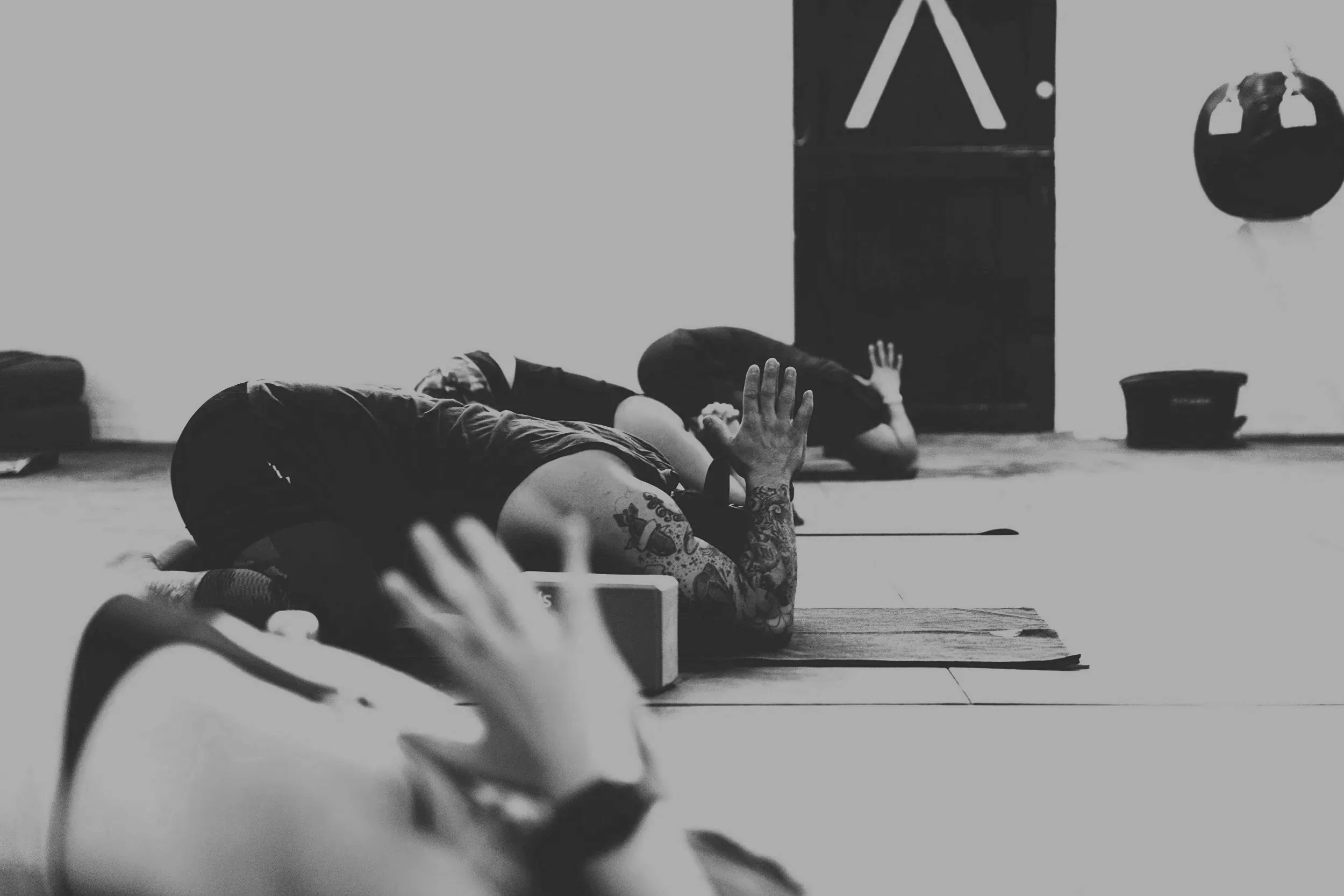 Deux personnes pratiquant la méditation ou le yoga en position allongée, dans une salle de classe ou un studio, avec un fond comprenant un tableau noir et une balle de fitness.