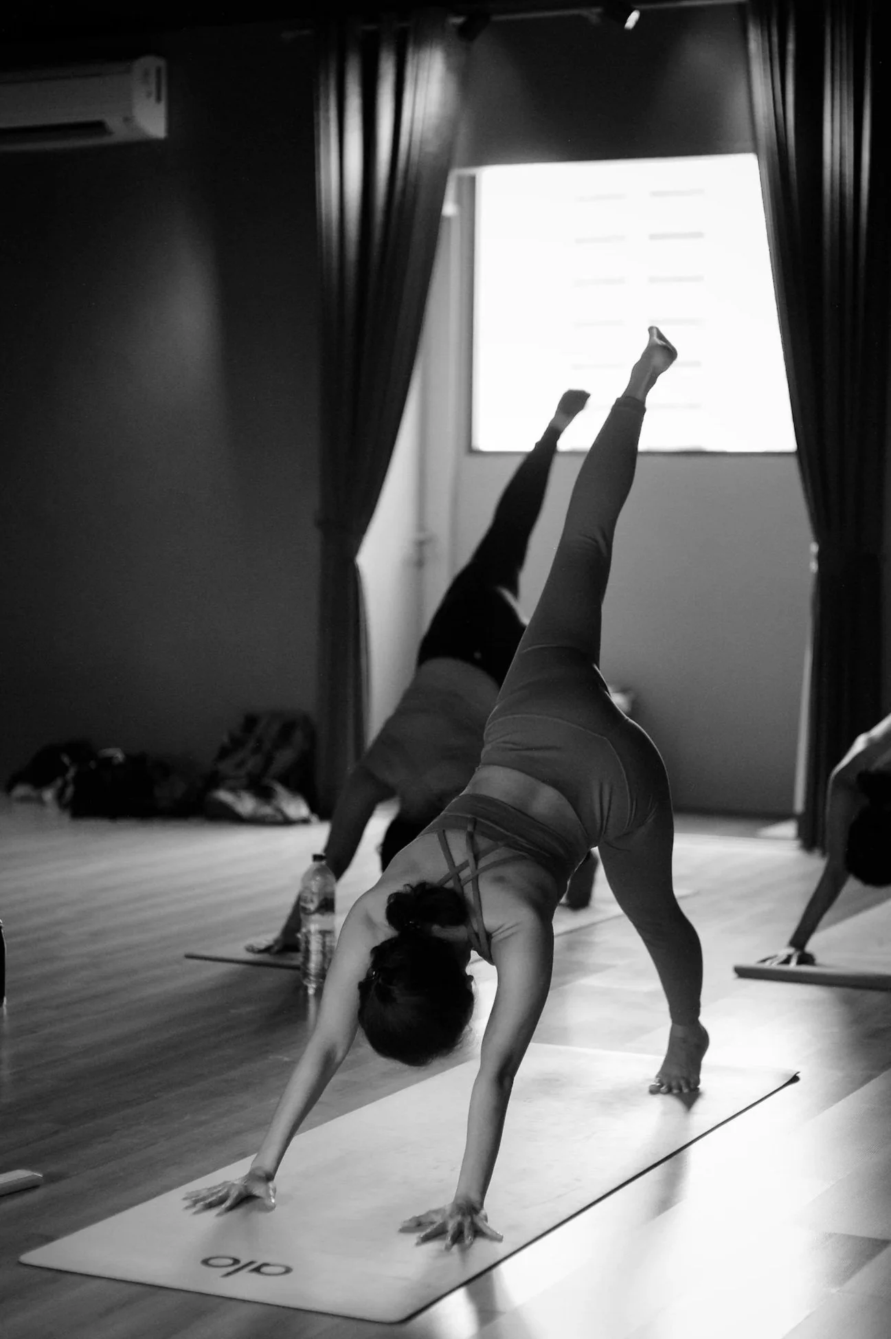 Une personne fait du yoga dans une pièce avec des rideaux noirs, un mur sombre, et une fenêtre avec de la lumière naturelle. Elle est en position de chien tête en bas sur un tapis de yoga.