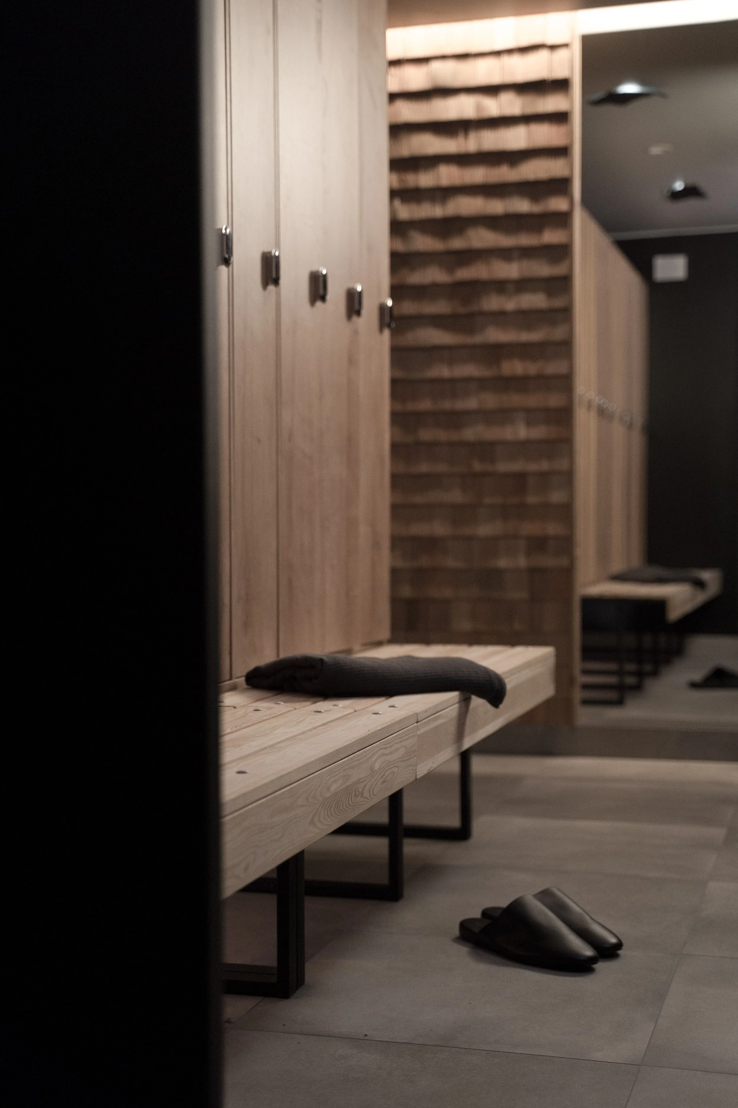 Une salle de vestiaire moderne avec des casiers en bois, un banc en bois, des pantoufles noires et un sol en carrelage gris.
