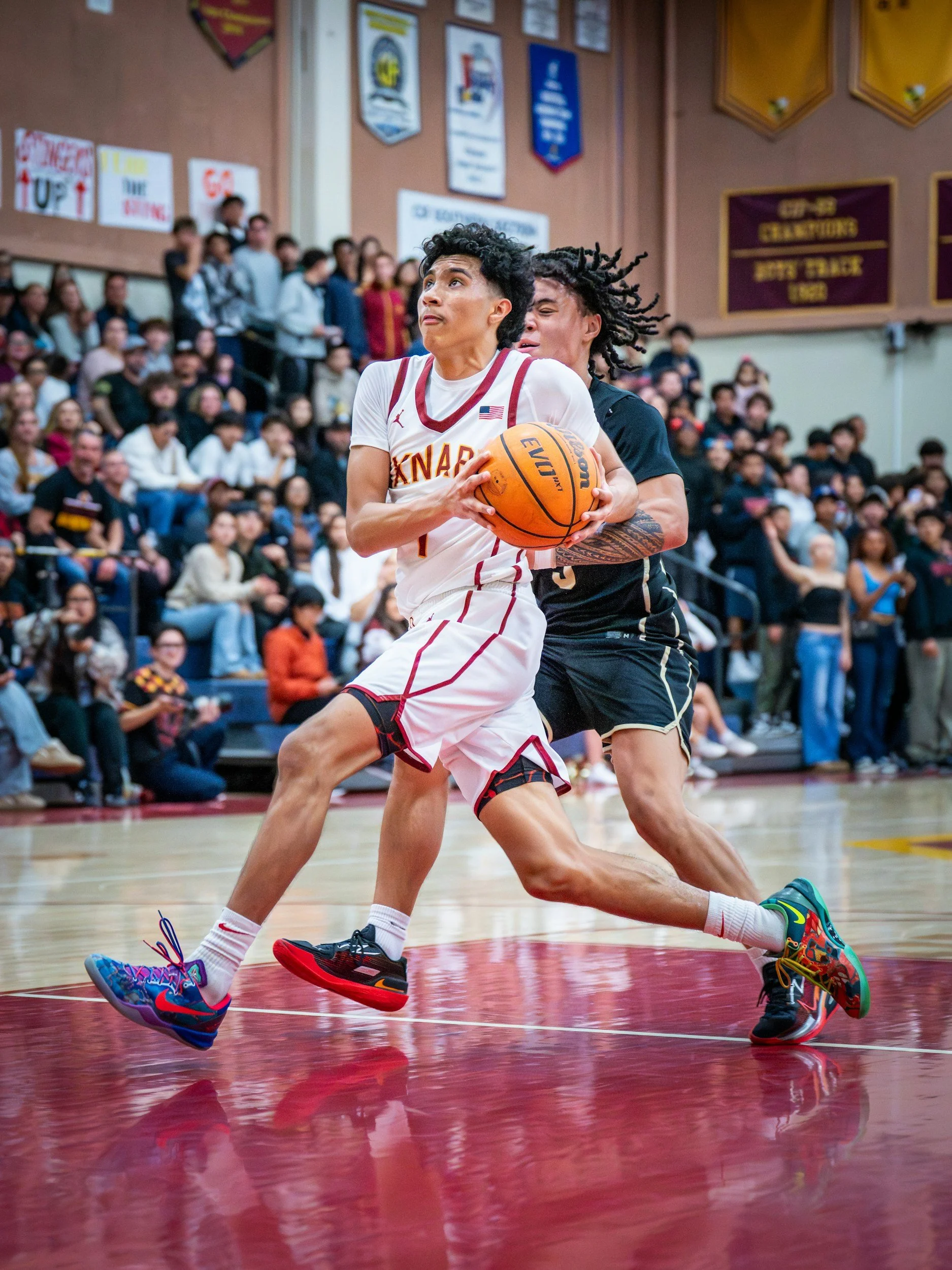 Two basketball players are competing for possession of the ball during a game in a gymnasium with spectators in the background. One player is in a white uniform with red accents, and the other in a black uniform. The player in white is holding the ball tightly while the player in black is reaching from behind. Both are wearing colorful sneakers.