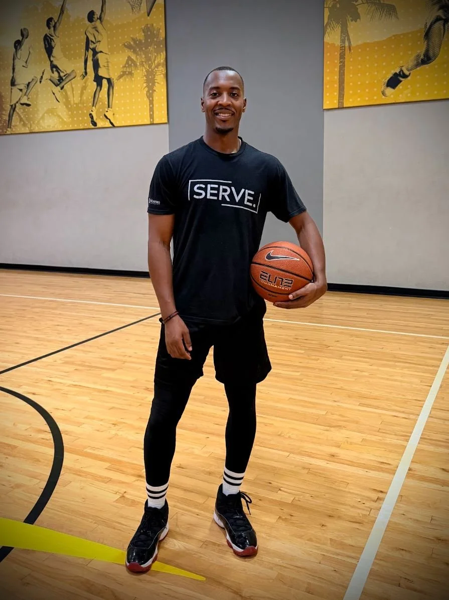 A basketball trainer is standing on an indoor basketball court holding a basketball, wearing a black t-shirt with the word 'SERVE' printed on it, smiling at the camera.