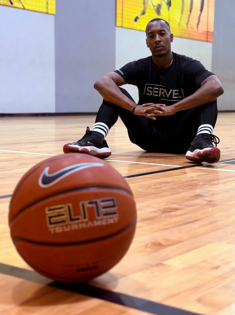 A basketball trainer sitting on a basketball court floor with a basketball in front of him, looking at the camera.
