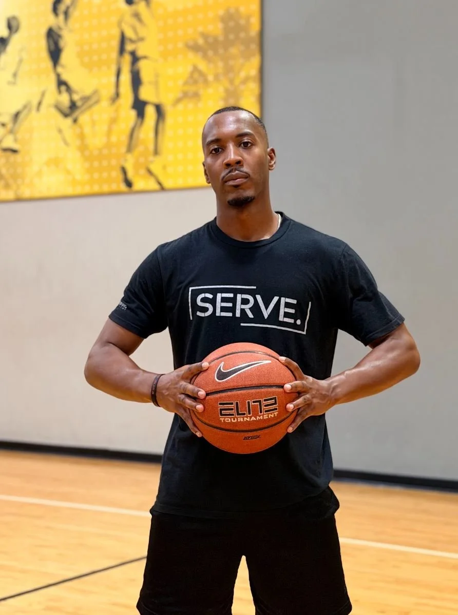 Basketball trainer holding a basketball stands on a basketball court wearing a 'SERVE' T-shirt