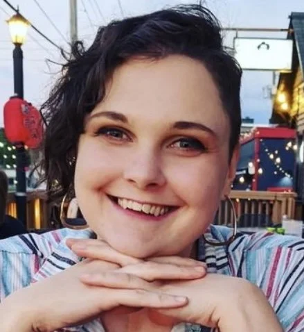 A smiling young woman with short, curly dark hair, sitting outdoors at a table with her hands under her chin. She is wearing a colorful striped top. The background includes decorative lights and a fenced area, suggesting an outdoor dining or entertainment setting.