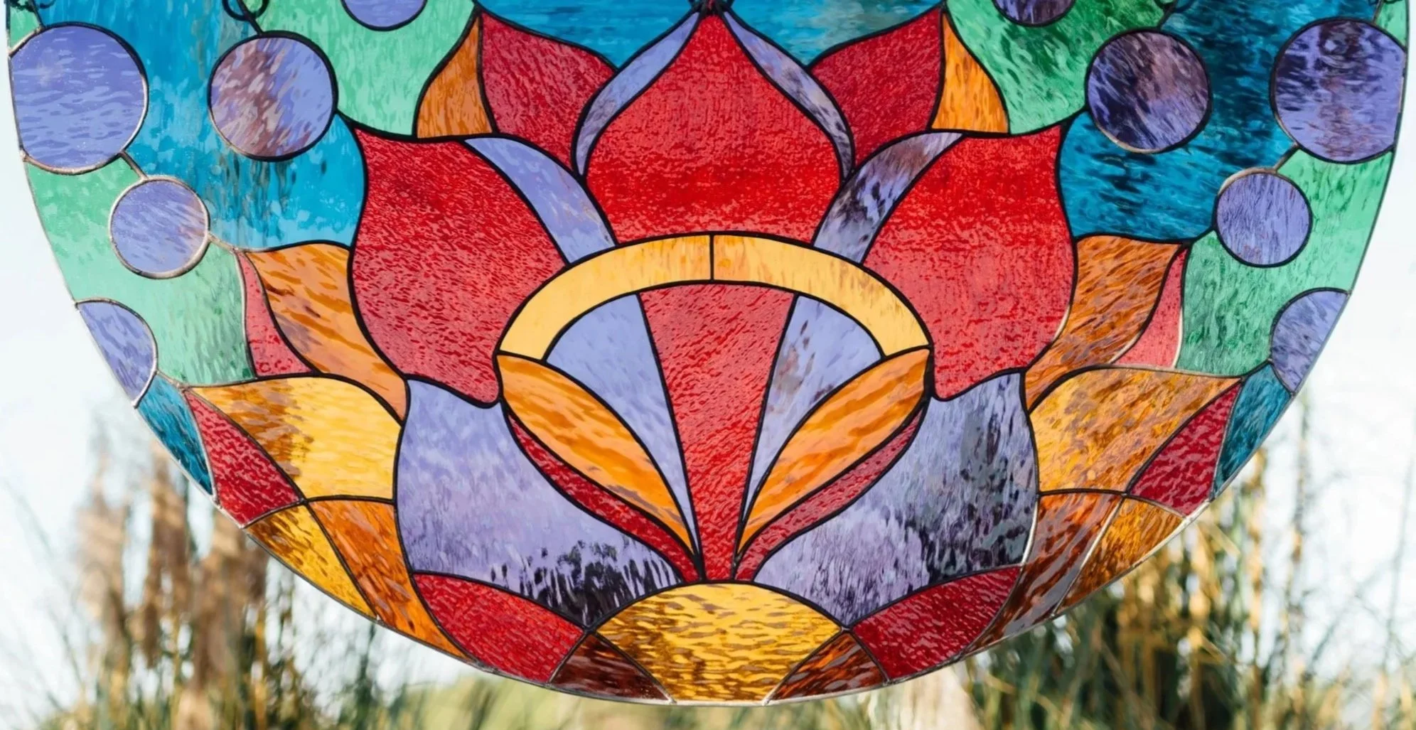 Colorful stained glass lampshade with intricate geometric patterns, hanging outdoors with trees in the blurred background.