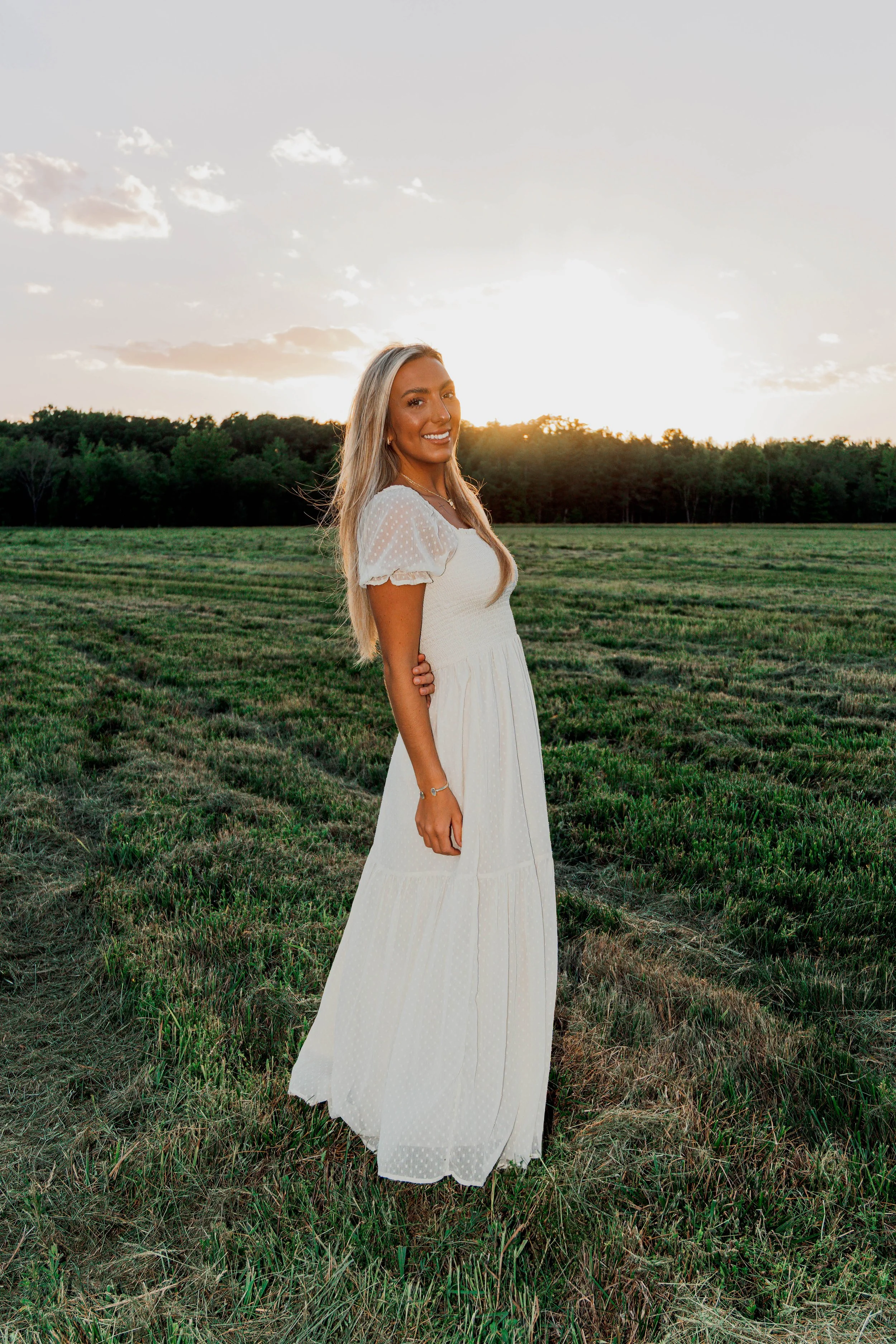 A woman with long blonde hair wearing a white dress standing in a grassy field at sunset with trees in the background.
