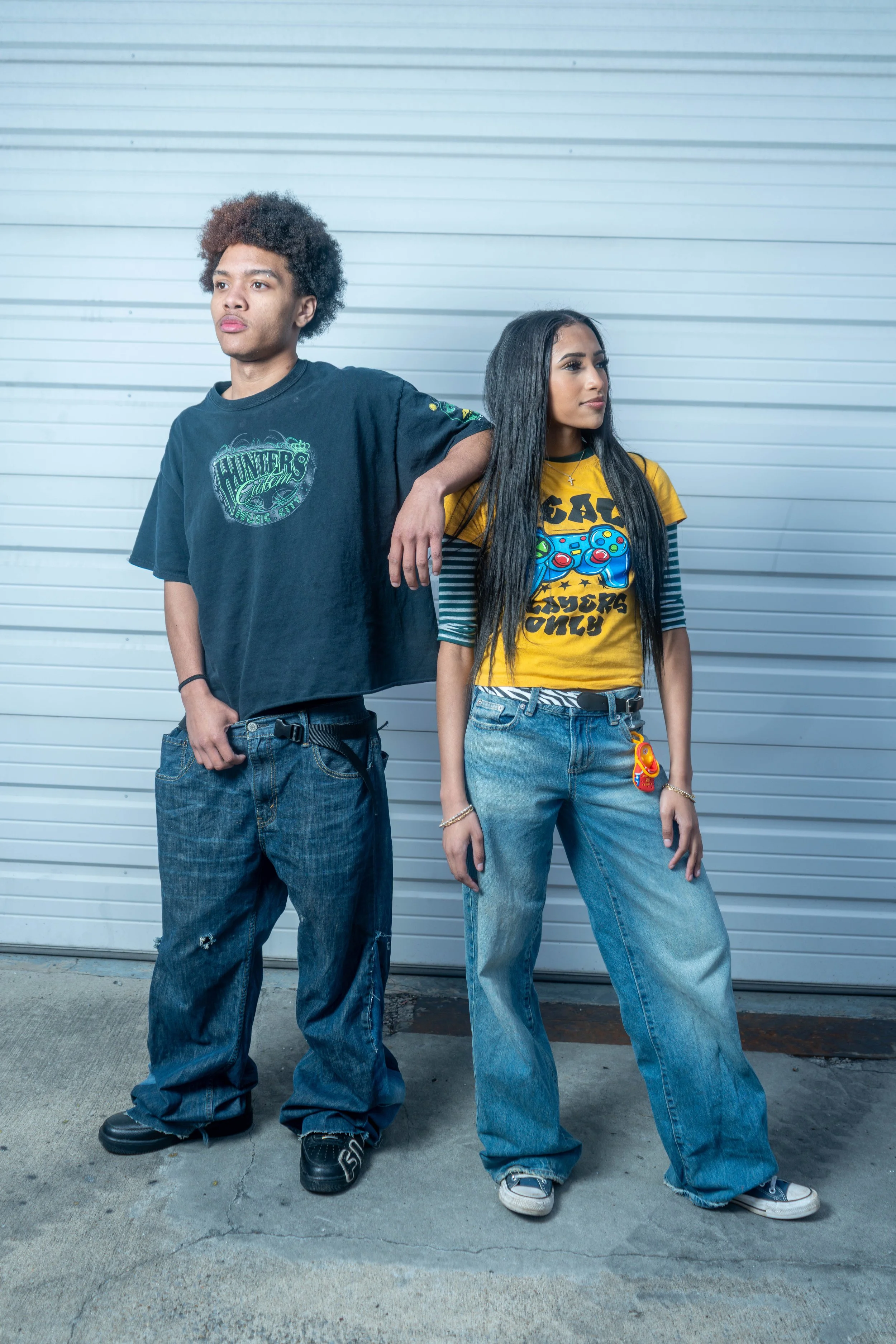 A young man with curly hair wearing a black T-shirt and baggy jeans stands next to a young woman with long black hair wearing a yellow graphic T-shirt and jeans, both leaning against a metal wall.