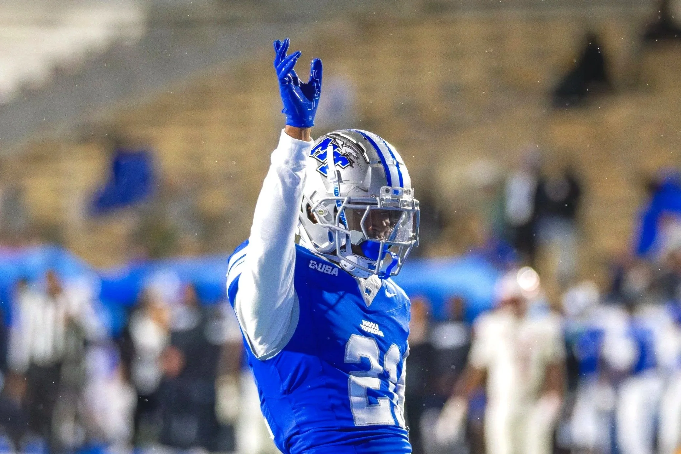 An American football player wearing a blue and white uniform with a helmet, extending his right arm upward with fingers in a gesture during a game on a field, with a blurred crowd in the background.