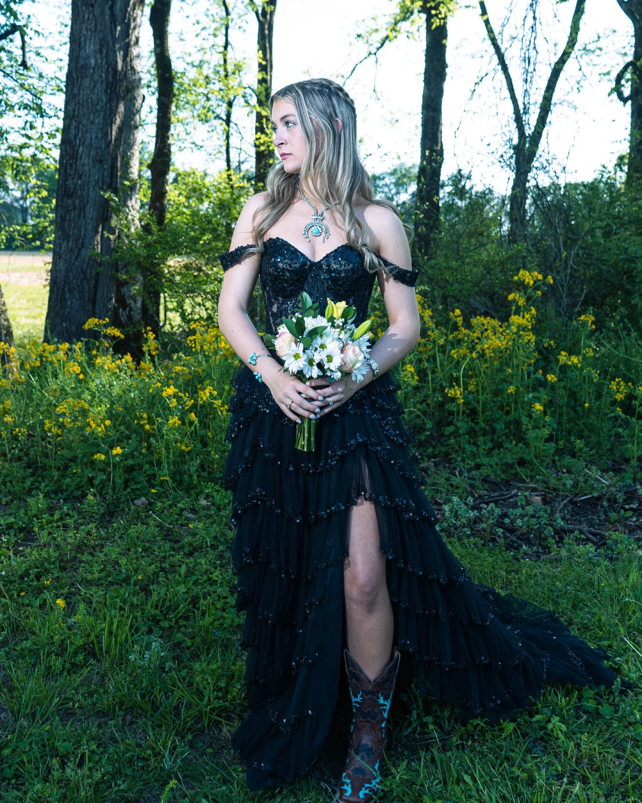 A woman in a black lace off-shoulder dress holding a bouquet of white and yellow flowers stands in a lush green forest with yellow wildflowers.