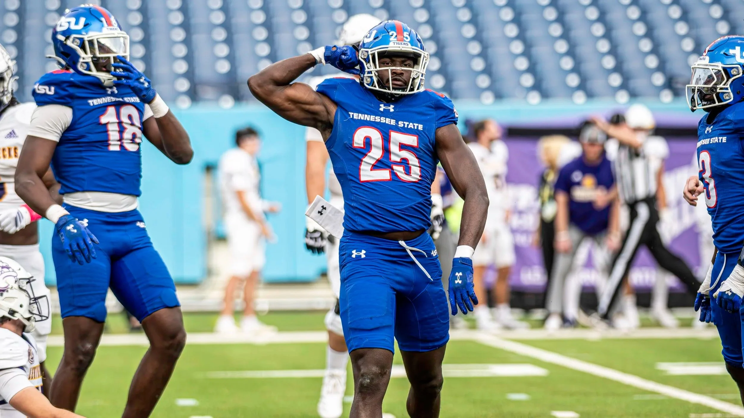 Tennessee State football players on the field during a game, with one player striking a pose by flexing his arm, all wearing blue uniforms and helmets.