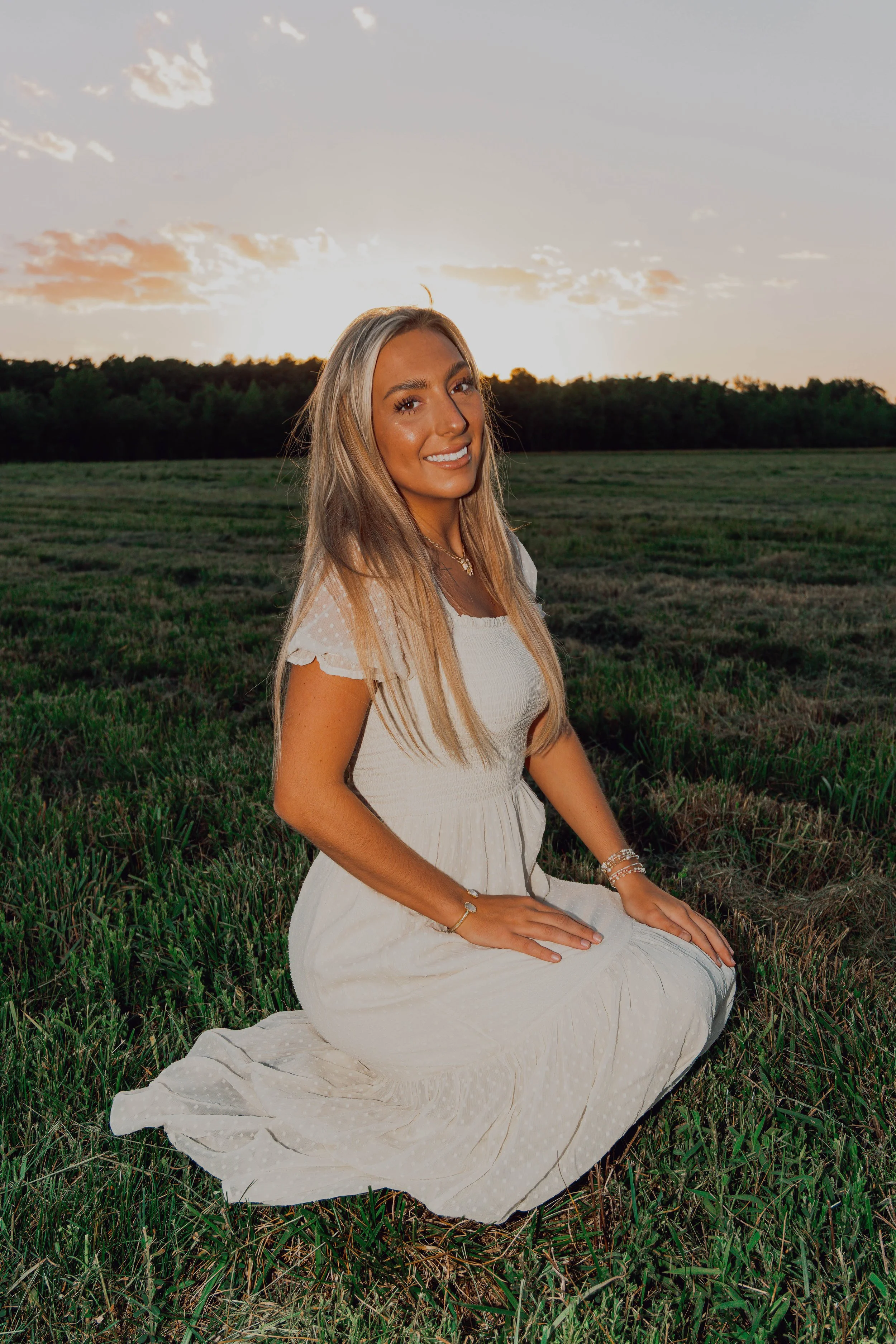 A woman in a white dress sitting on grass during sunset, smiling at the camera.