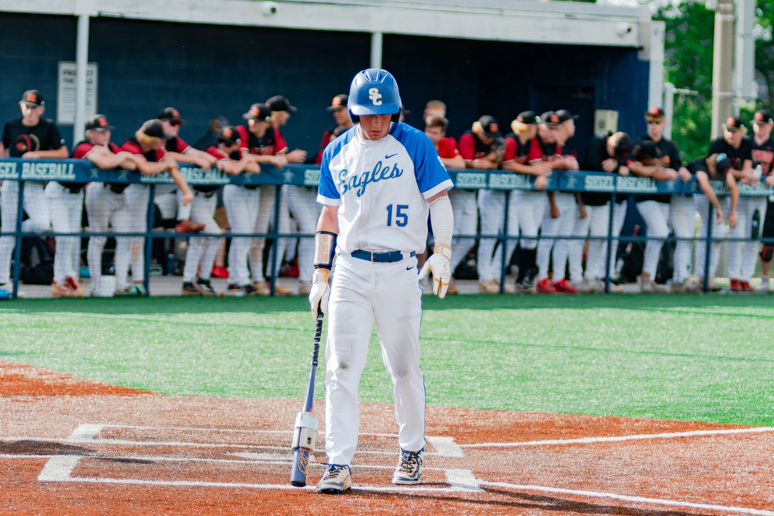 Baseball player in white and blue uniform standing at home plate with a bat, in front of a dugout with team members in black and red uniforms leaning over the fence.