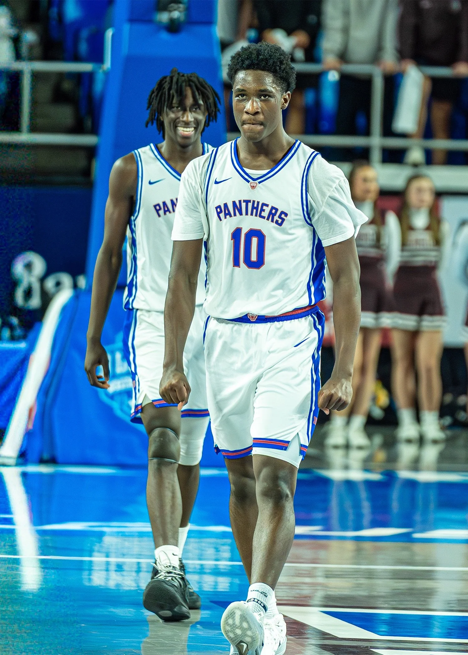 Two basketball players in white jerseys with blue trim, one wearing number 10, walking on a basketball court. The player in front has a focused expression, while the player behind is smiling. Spectators are visible in the background.