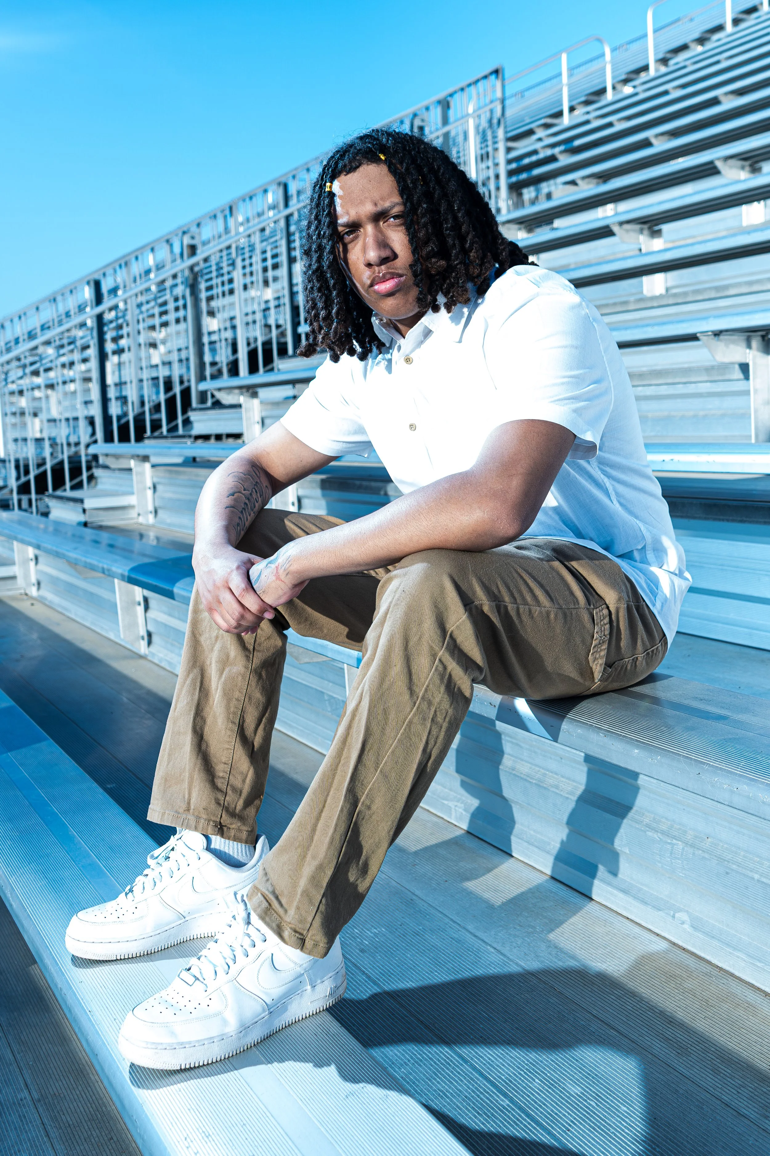 A young man with dreadlocks sitting on metal bleachers outdoors under a clear blue sky, wearing a white shirt, brown pants, and white sneakers.