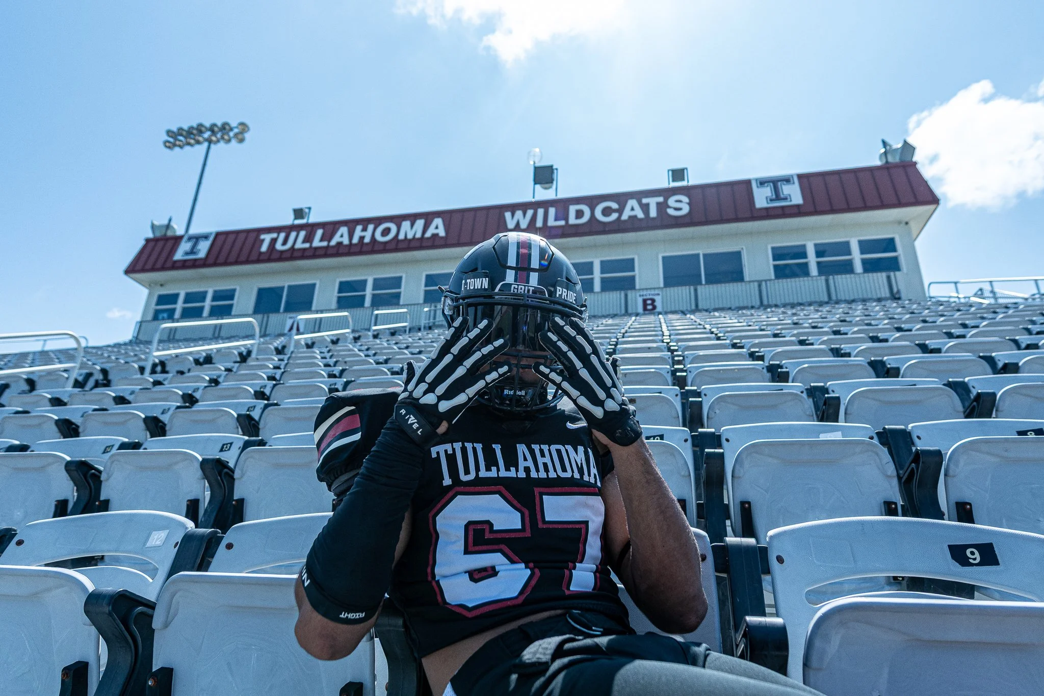Person in a Tullahoma Wildcats football uniform and helmet sitting in empty stadium seats, holding their helmet with both hands covering their face.