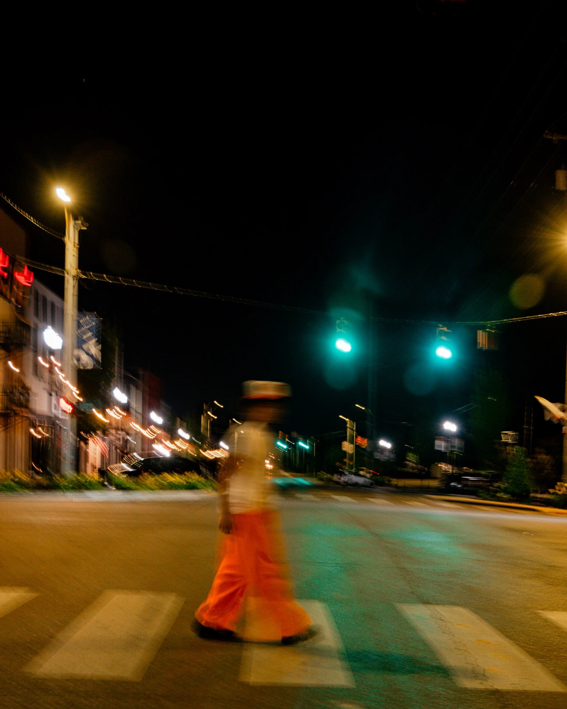 Nighttime street scene with a person in an orange outfit walking in a crosswalk, blurred lights, buildings, and traffic signals.