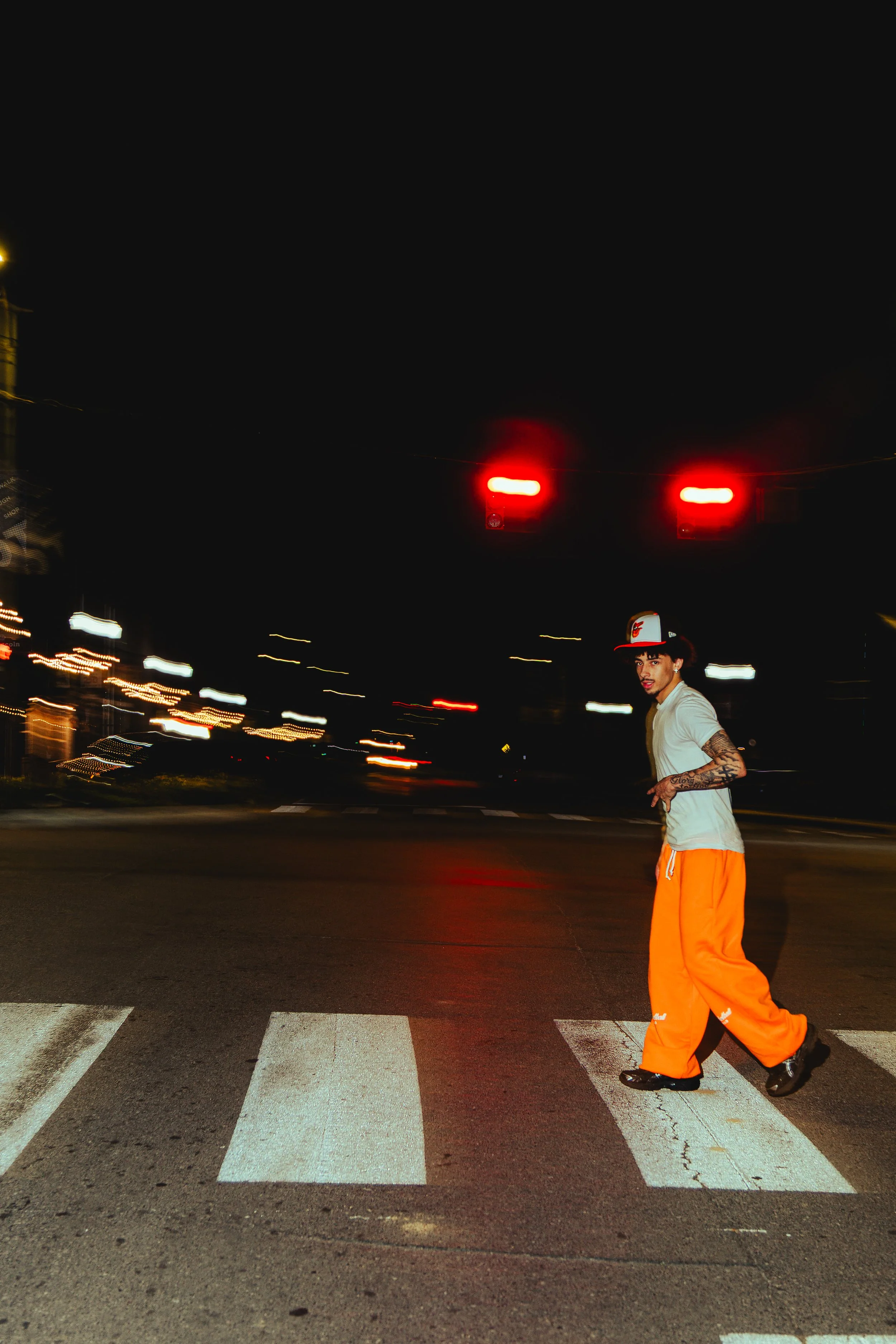 A young man wearing orange pants, a white shirt, sneakers, and a cap crossing a street at night in front of a traffic light showing red, with blurred city lights in the background.