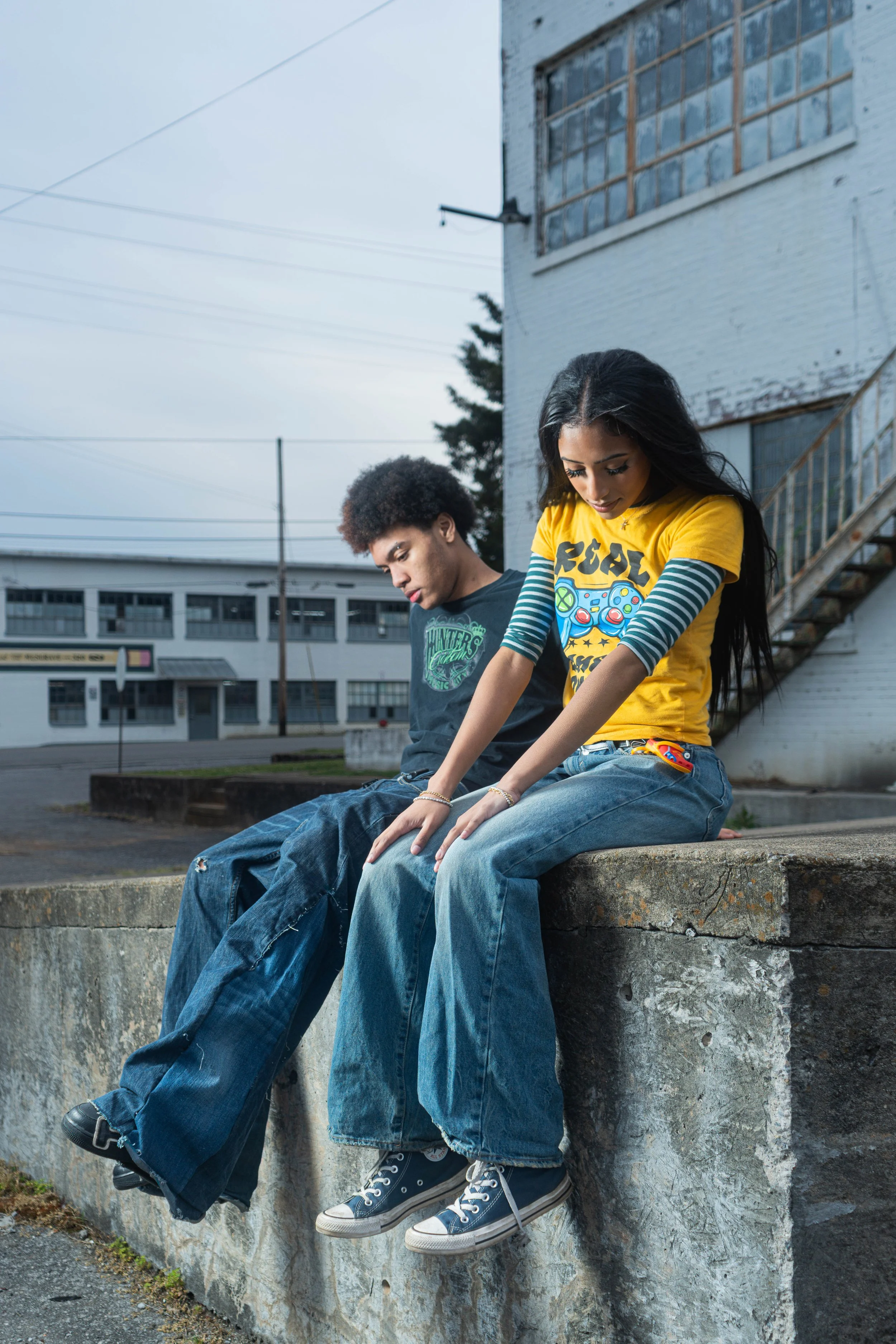 Two young people sitting on a concrete ledge outdoors, with a building and utility poles in the background. The person on the right has long dark hair, is wearing a yellow graphic t-shirt and jeans, and is looking down. The person on the left has curly hair, is wearing a dark t-shirt and jeans, and is also looking down.