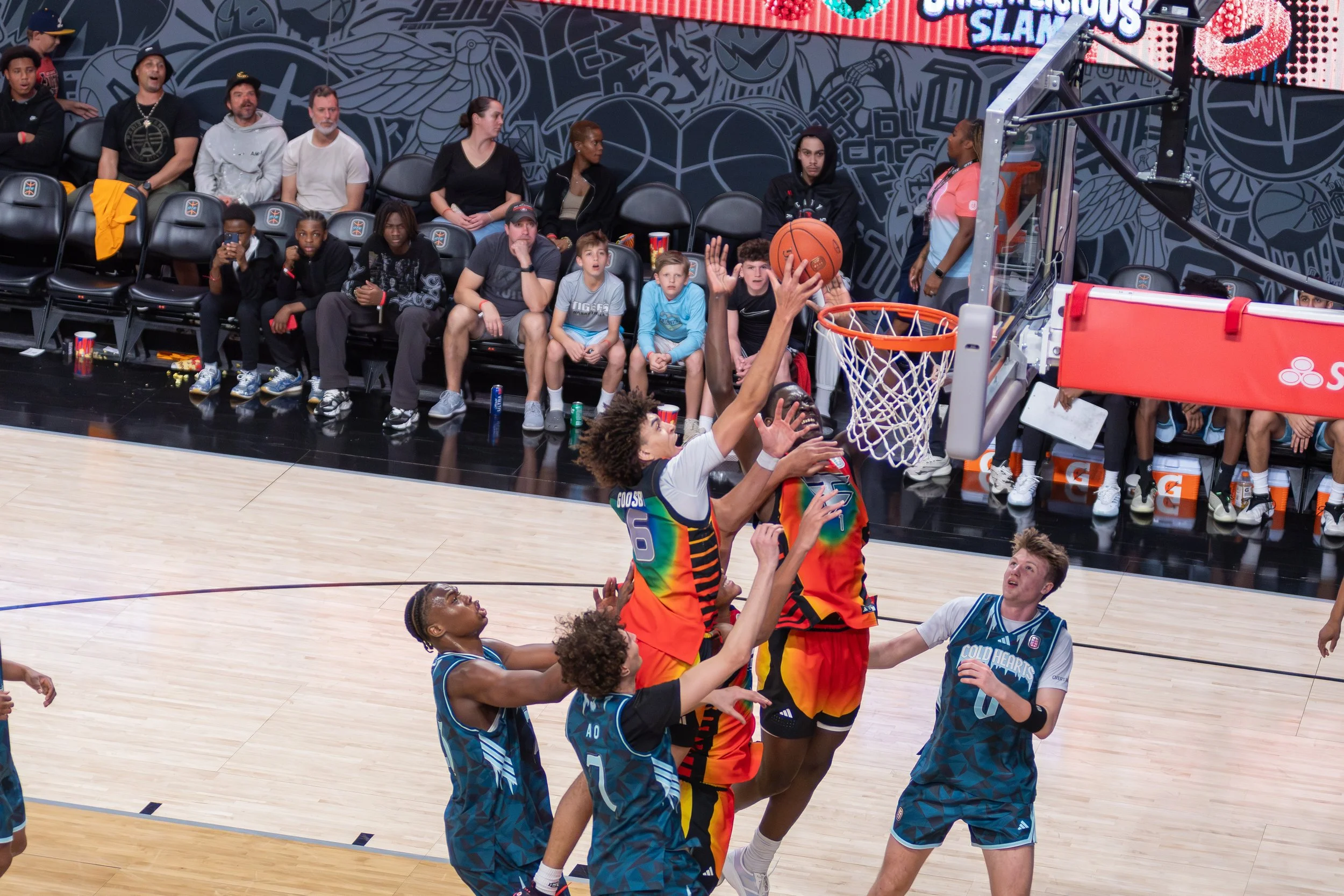 A basketball game with players reaching for the ball near the hoop, and spectators sitting courtside watching the game.