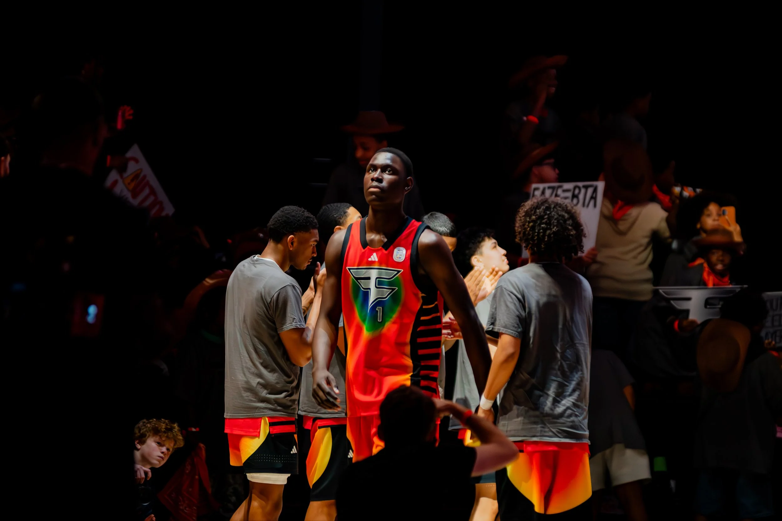 A basketball player in a colorful jersey stands on the court, surrounded by spectators and teammates, with a dark background and some people holding signs.