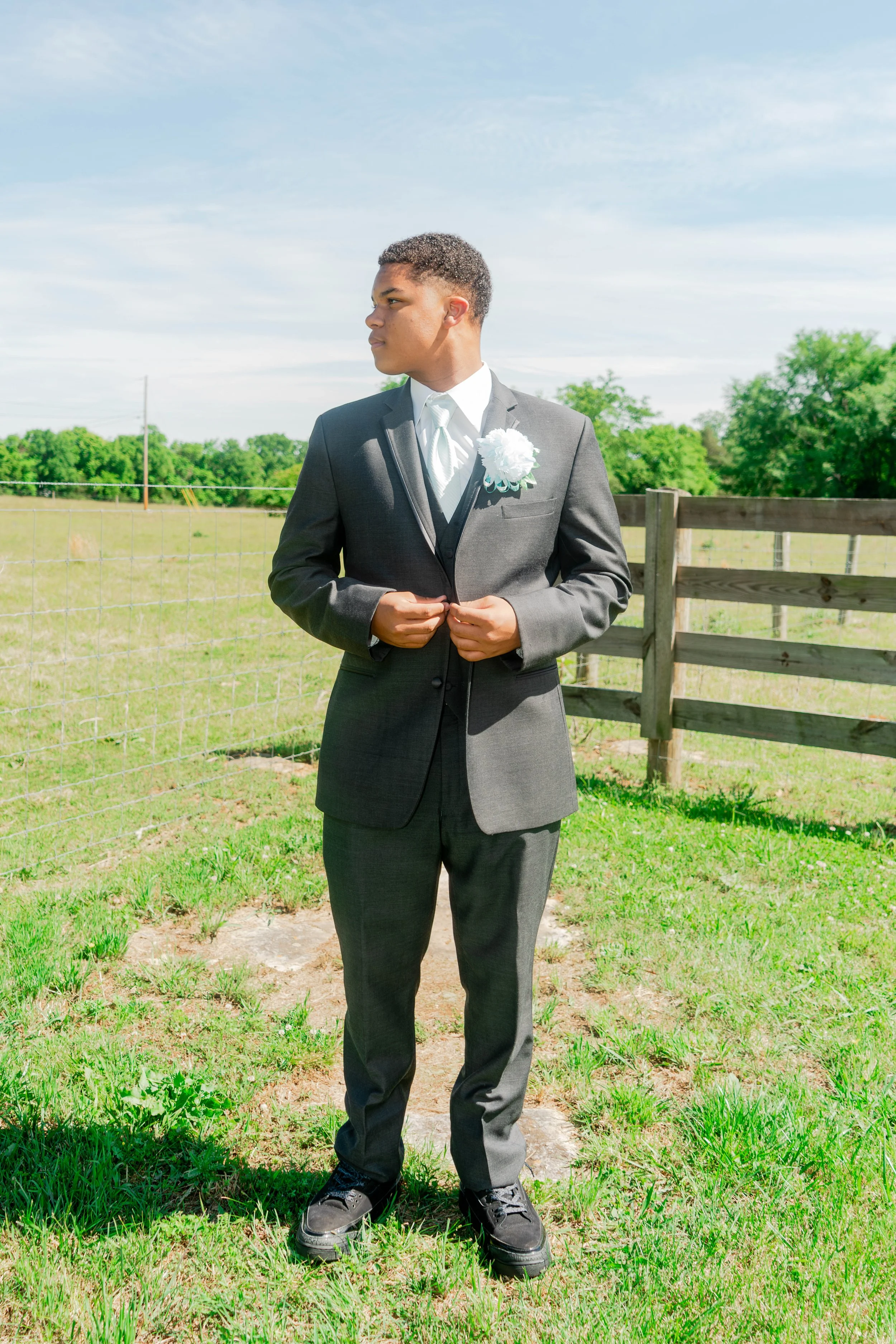 A young man in a gray suit and tie stands outdoors in a green field with trees and a wooden fence behind him, looking to his left.