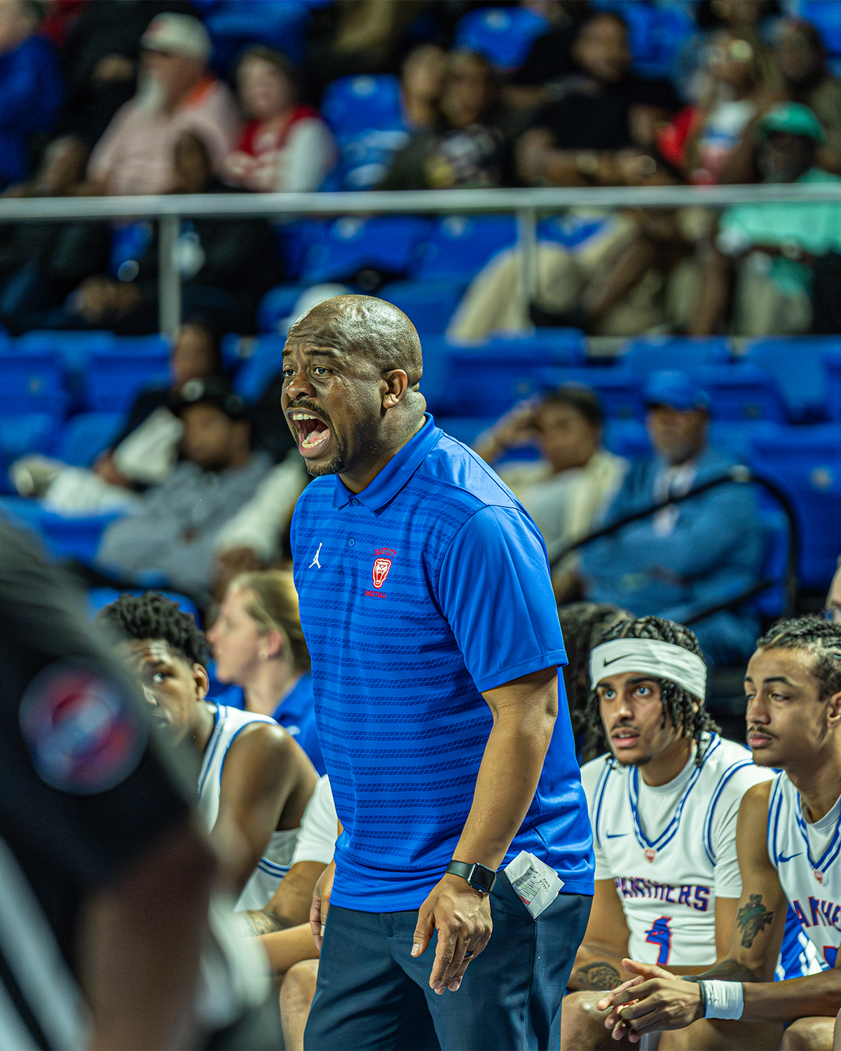 A basketball coach yelling on the sideline during a game, wearing a blue shirt with a team logo, surrounded by players sitting on the bench in basketball jerseys.