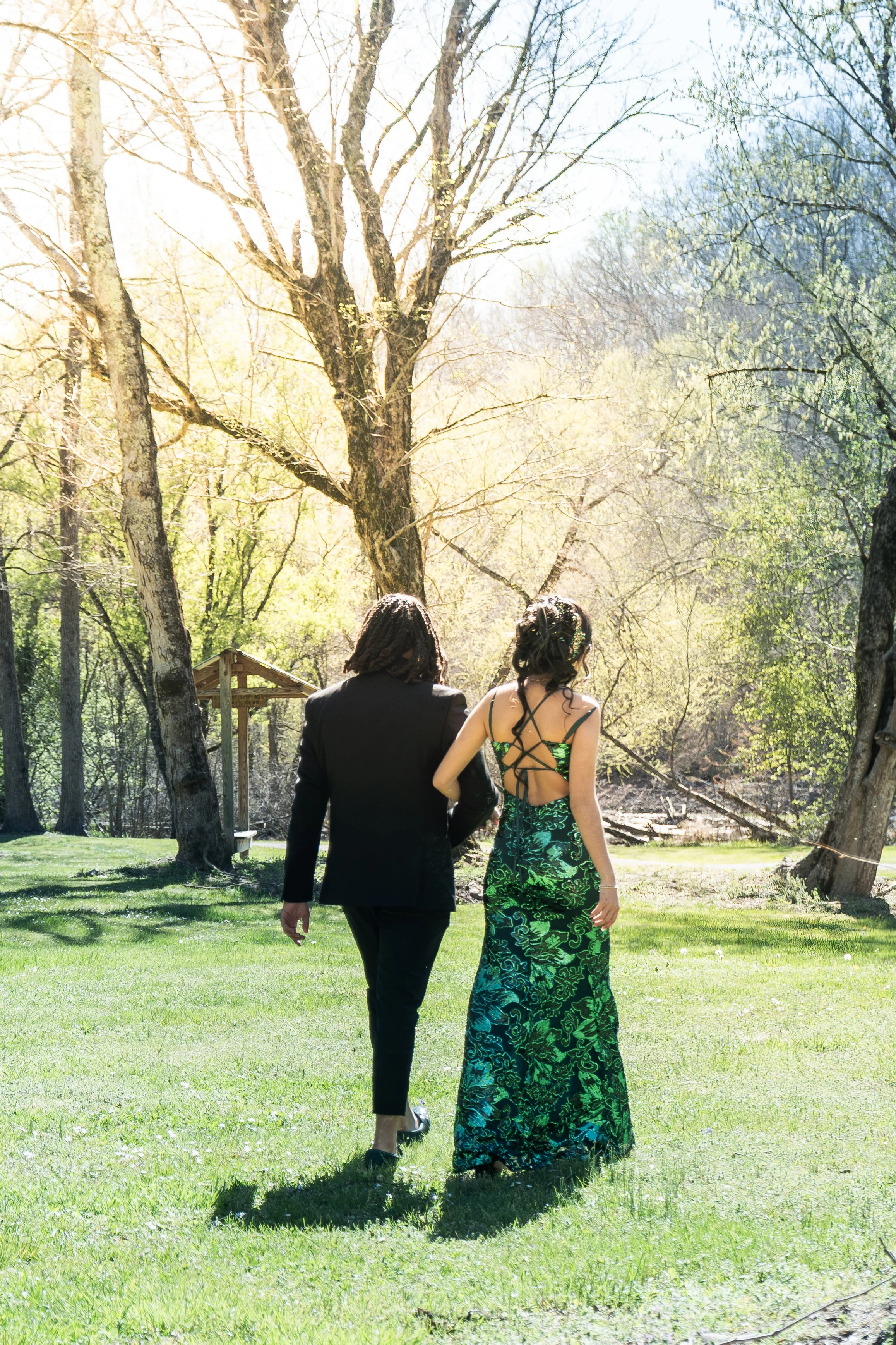 A couple dressed in formal attire walking in a grassy park with large leafless trees in the background on a sunny day.