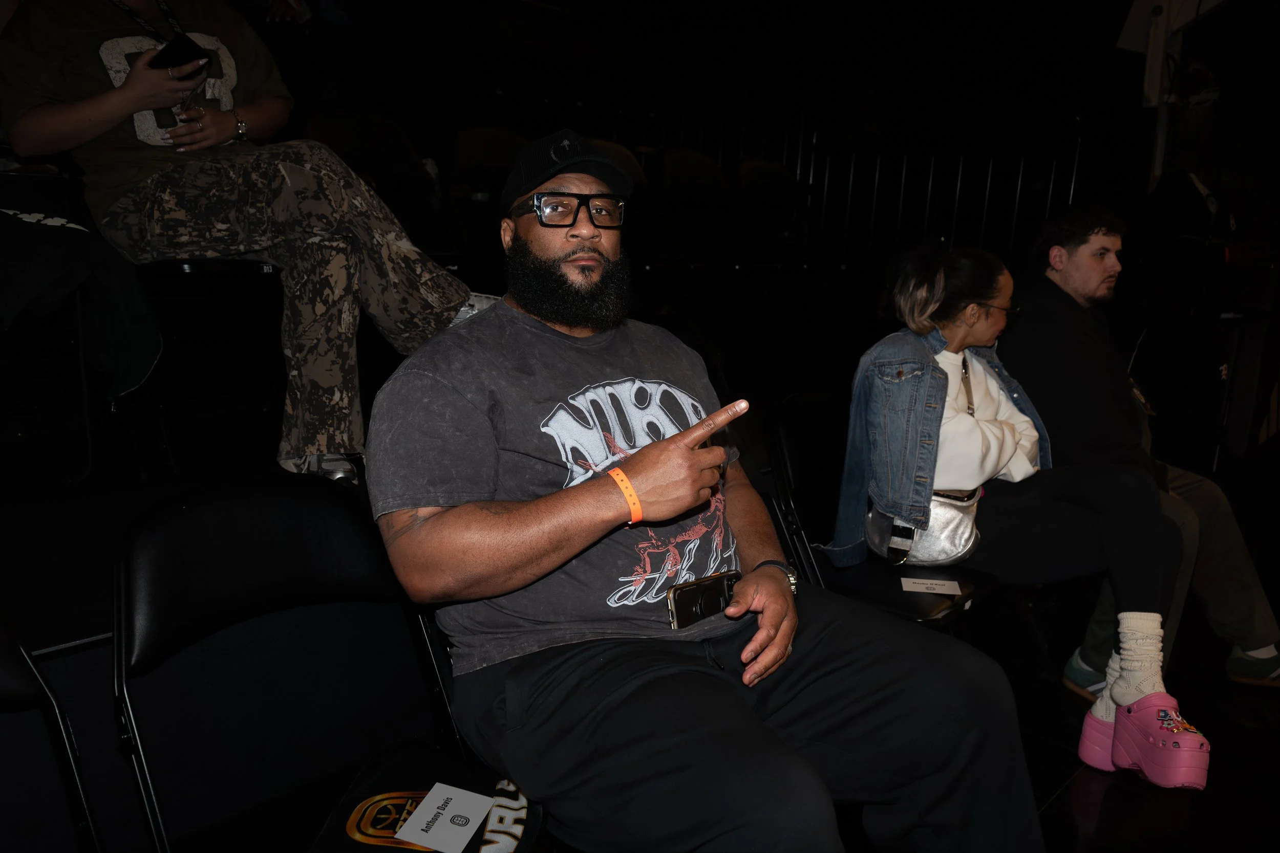 A man with a beard and glasses sitting in a dark room, making a peace sign with his right hand. He is wearing a black cap, a graphic T-shirt, and has a phone in his left hand. There are other people sitting nearby, including a woman in sunglasses and a denim jacket and a young man with curly hair.