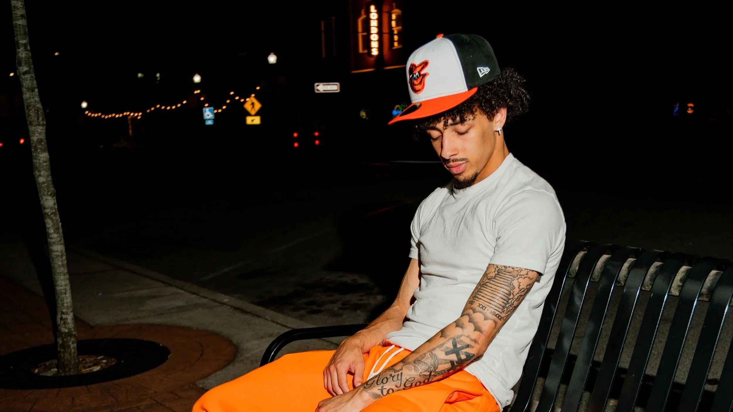A young man with curly hair and tattoos on his arms sitting on a bench at night with his eyes closed, wearing an orange and white Baltimore Orioles baseball cap and bright orange pants.