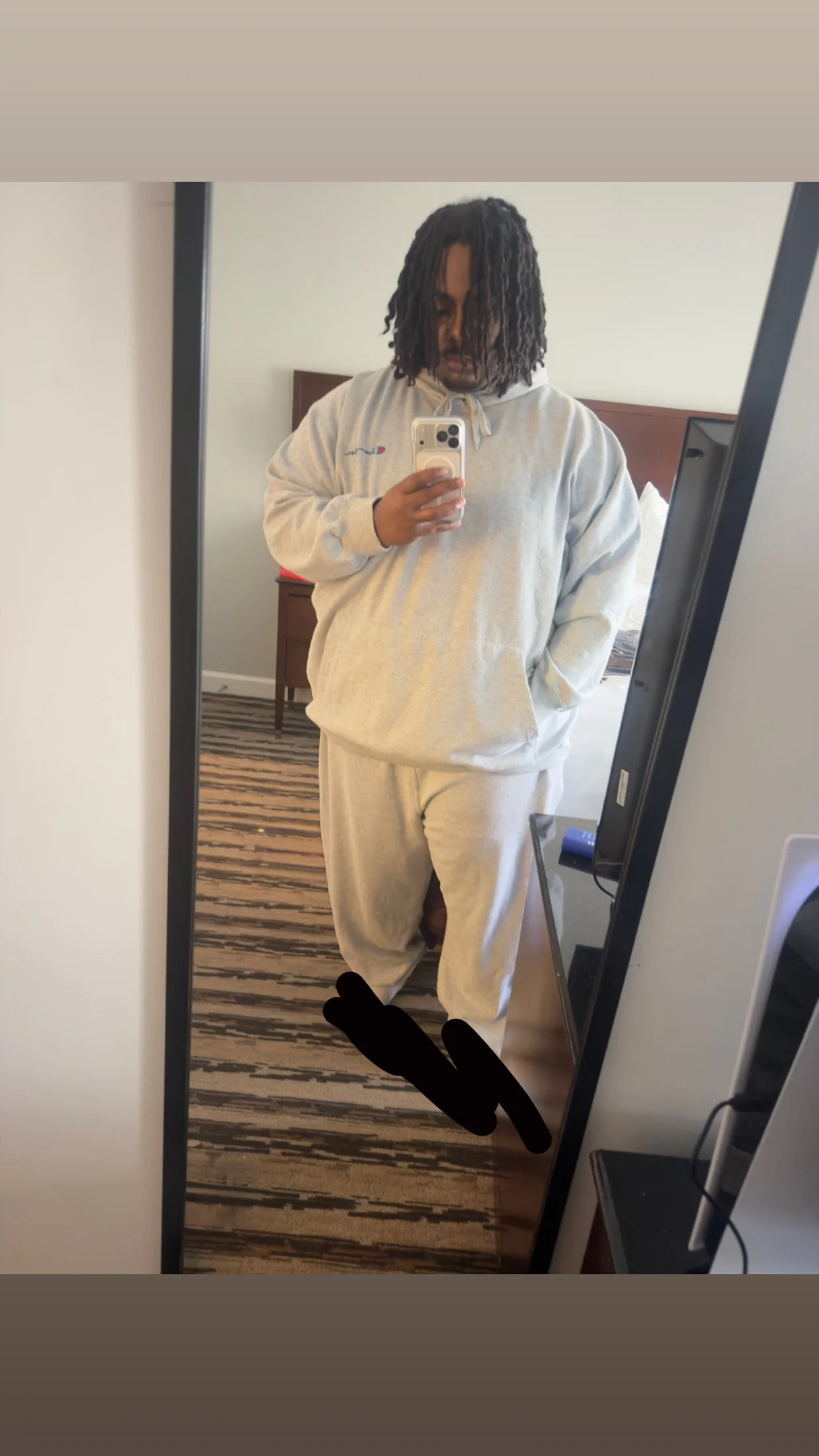 Person taking a mirror selfie in a hotel room, wearing a light gray hoodie and sweatpants, with dreadlocked hair partially covering face.