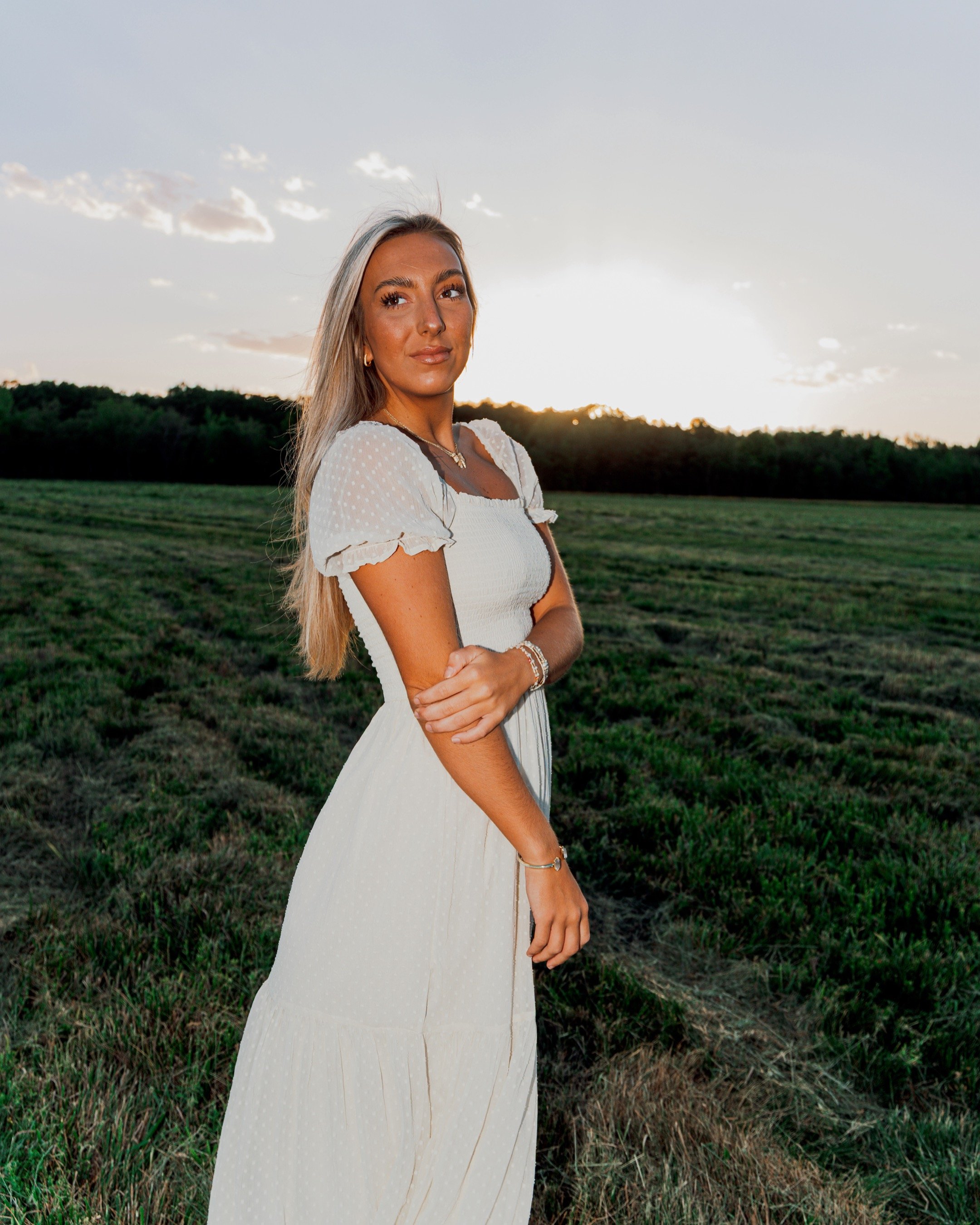 A woman with long blonde hair, wearing a white dress and jewelry, stands in a field at sunset, with a background of clouds and trees.