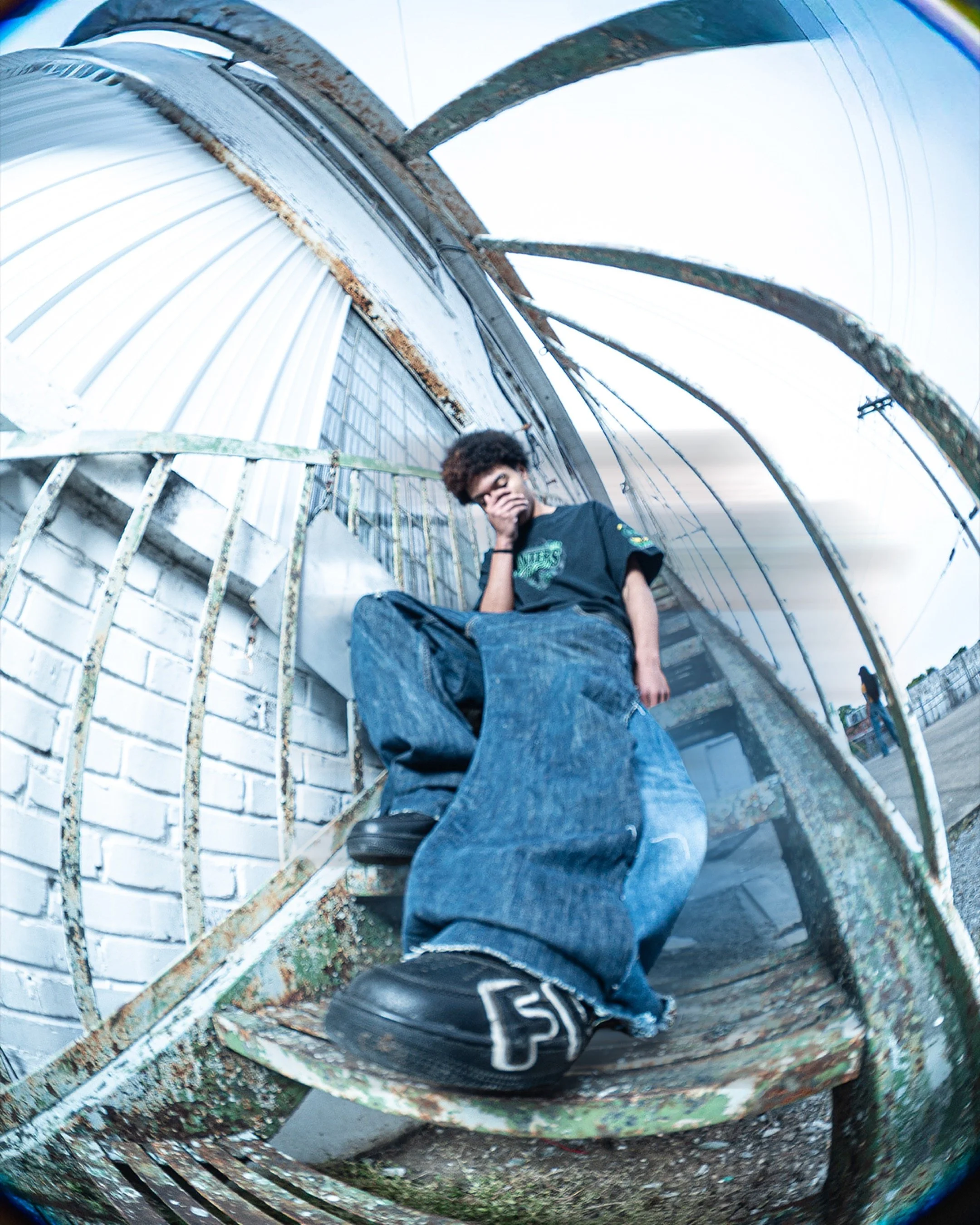 A person sitting inside a rusty, graffiti-covered, dome-shaped structure, with a hand covering their face, wearing a dark shirt and jeans, surrounded by an industrial outdoor setting.