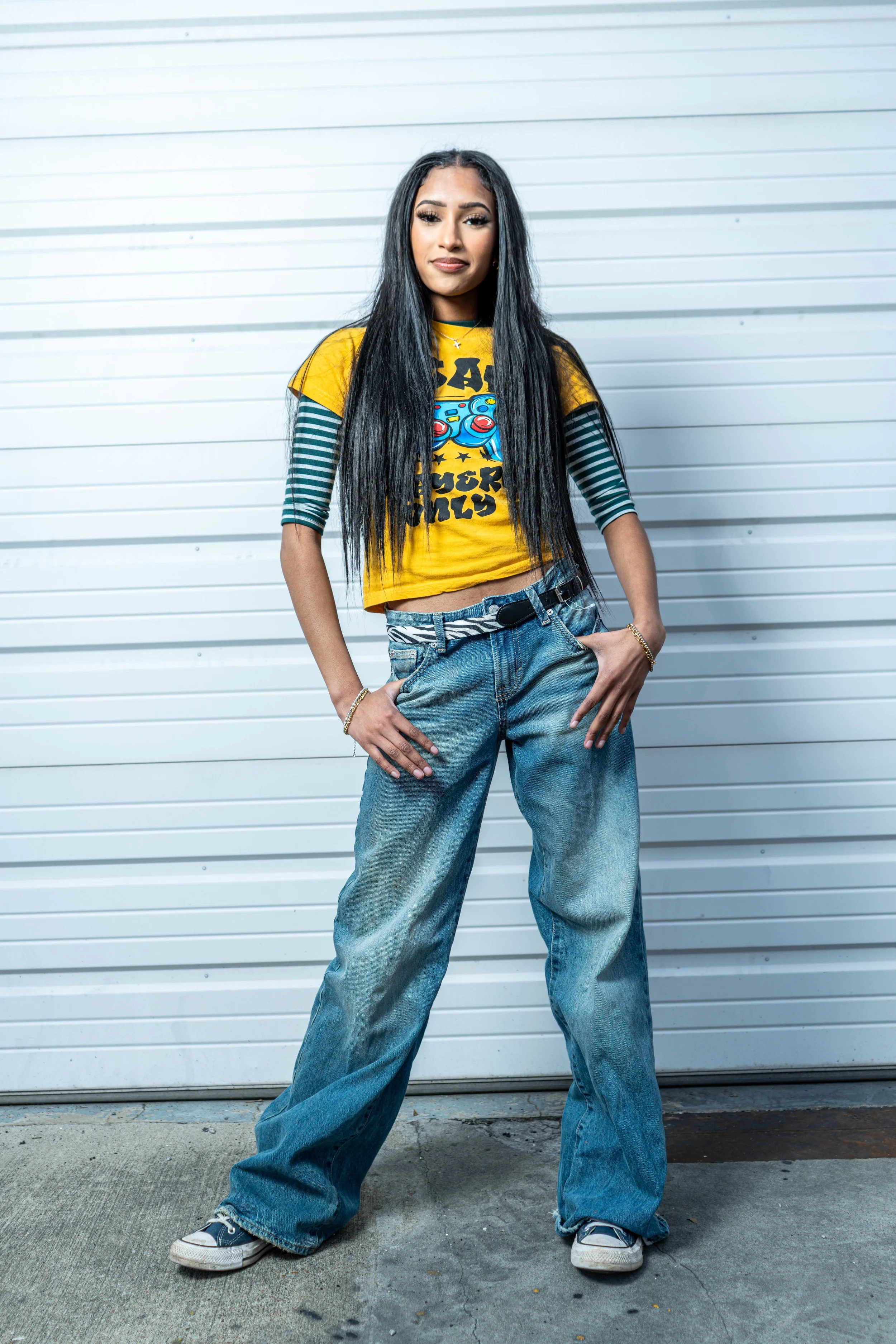 Young woman with long black hair standing in front of a white metal garage door, wearing a yellow graphic t-shirt layered over a striped long-sleeve shirt, baggy jeans, and sneakers, with her hands in her pockets.