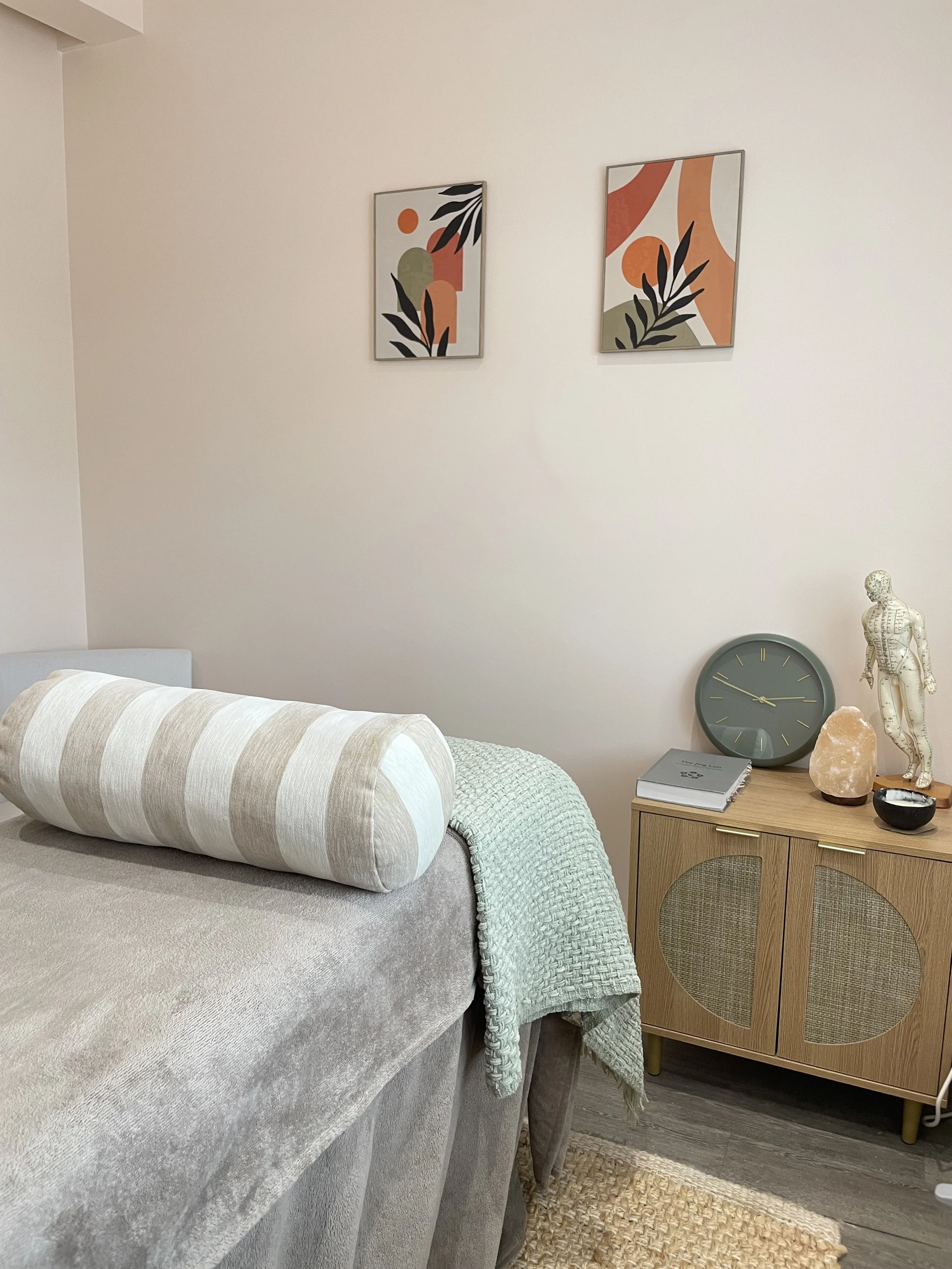 Living room with a beige sofa, decorative pillows, wall art with abstract designs, a wooden cabinet with decor items including a salt lamp, a clock, a dinosaur figurine, and a book, all in a neutral color palette.