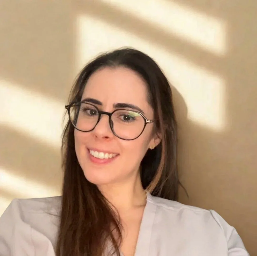 A smiling woman with long brown hair and glasses taking a selfie against a beige wall with sunlight creating shadow patterns.