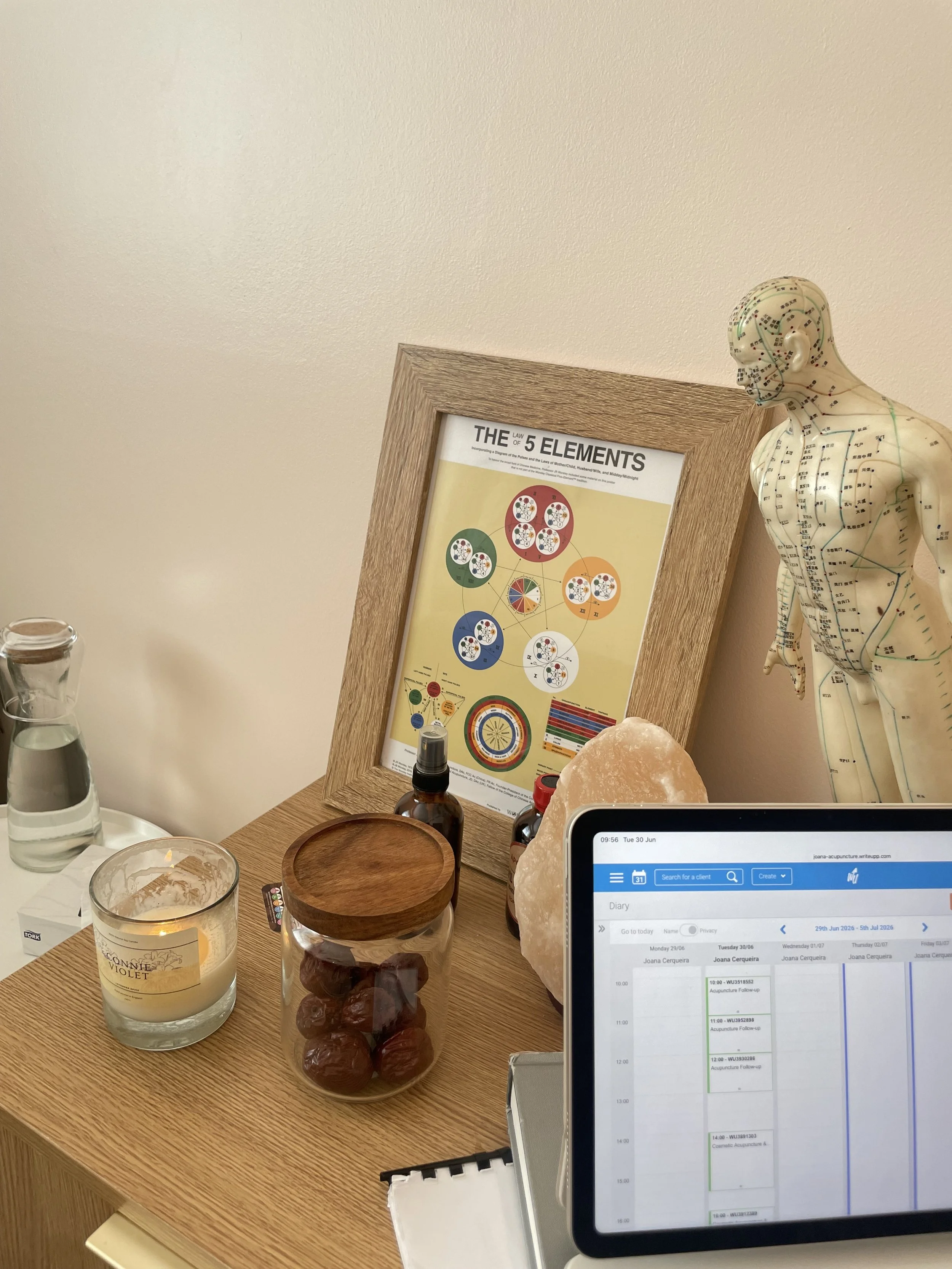A desk with candles, a small wooden bowl, a glass jar filled with dark brown objects, a salt lamp, a bottle of essential oil, an eye chart, a framed chart titled "The 5 Elements", and a tablet displaying a calendar and schedule. To the right, there is a human body diagram model with acupuncture points.