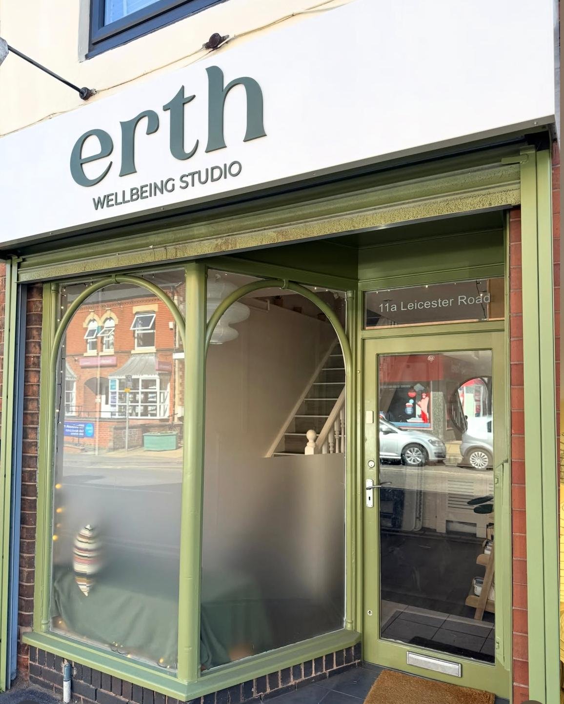 Green storefront with a sign that reads 'ertn WELLBEING STUDIO' and an address '11a Leicester Road', showing glass windows and a door with reflections of parked cars and buildings across the street.