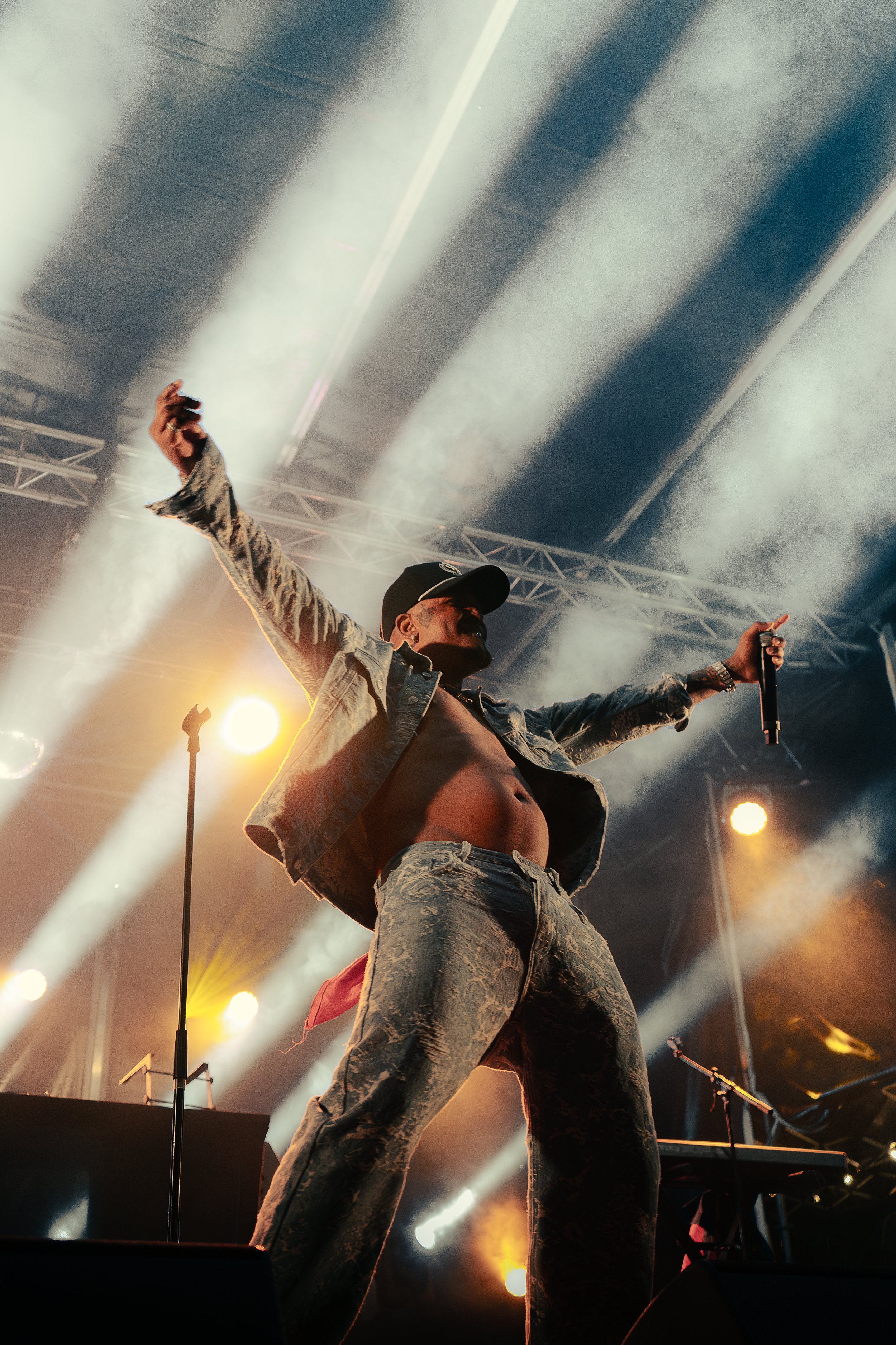 Un homme chante sur scène lors d'un concert, avec des des lumières colorées en arrière-plan, portant une veste en jean et un pantalon assorti, avec un chapeau de baseball.