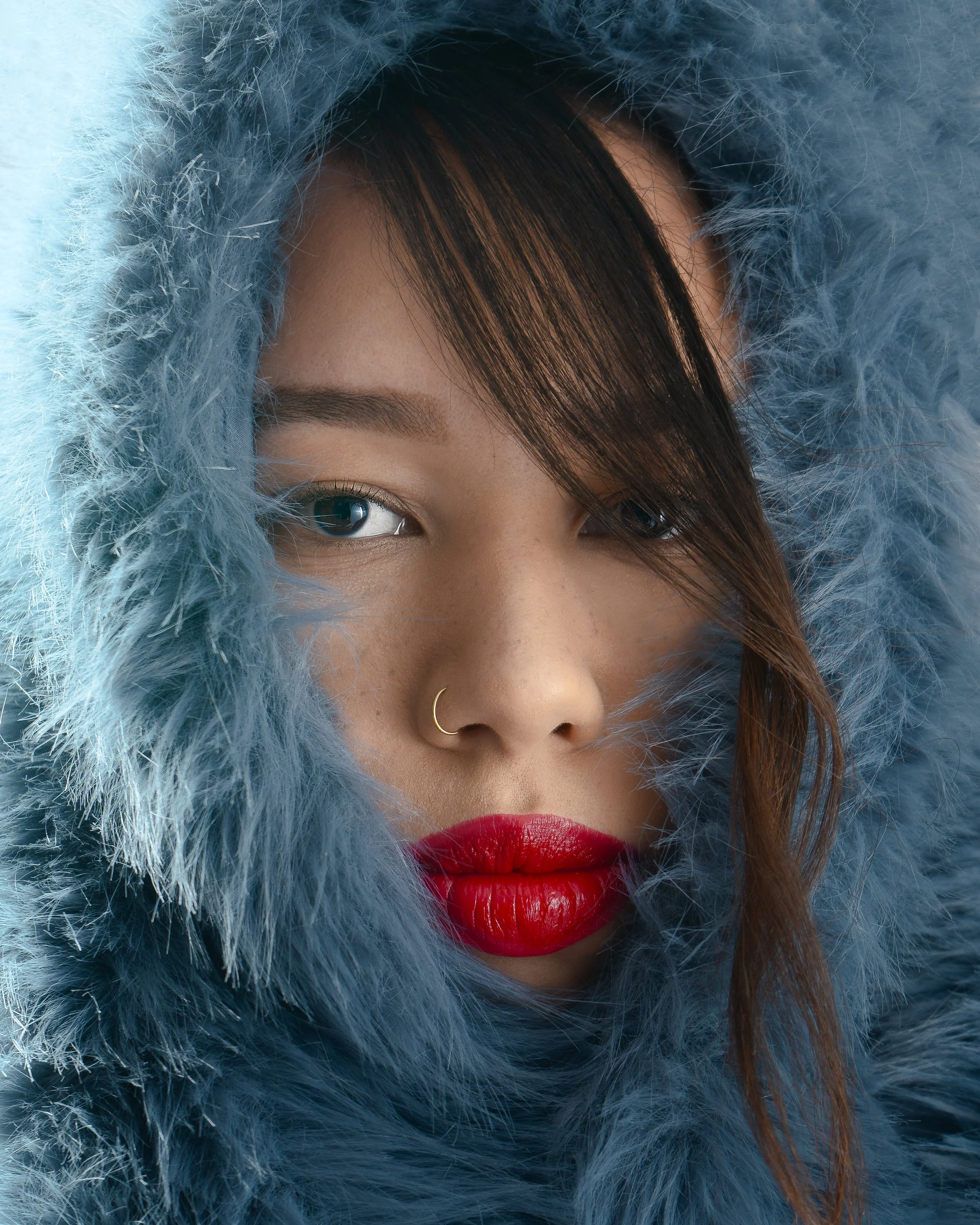 Une femme portant une capuche en fausse fourrure bleue, avec du maquillage rouge sur les lèvres, un anneau nasal en or, et des yeux bleus