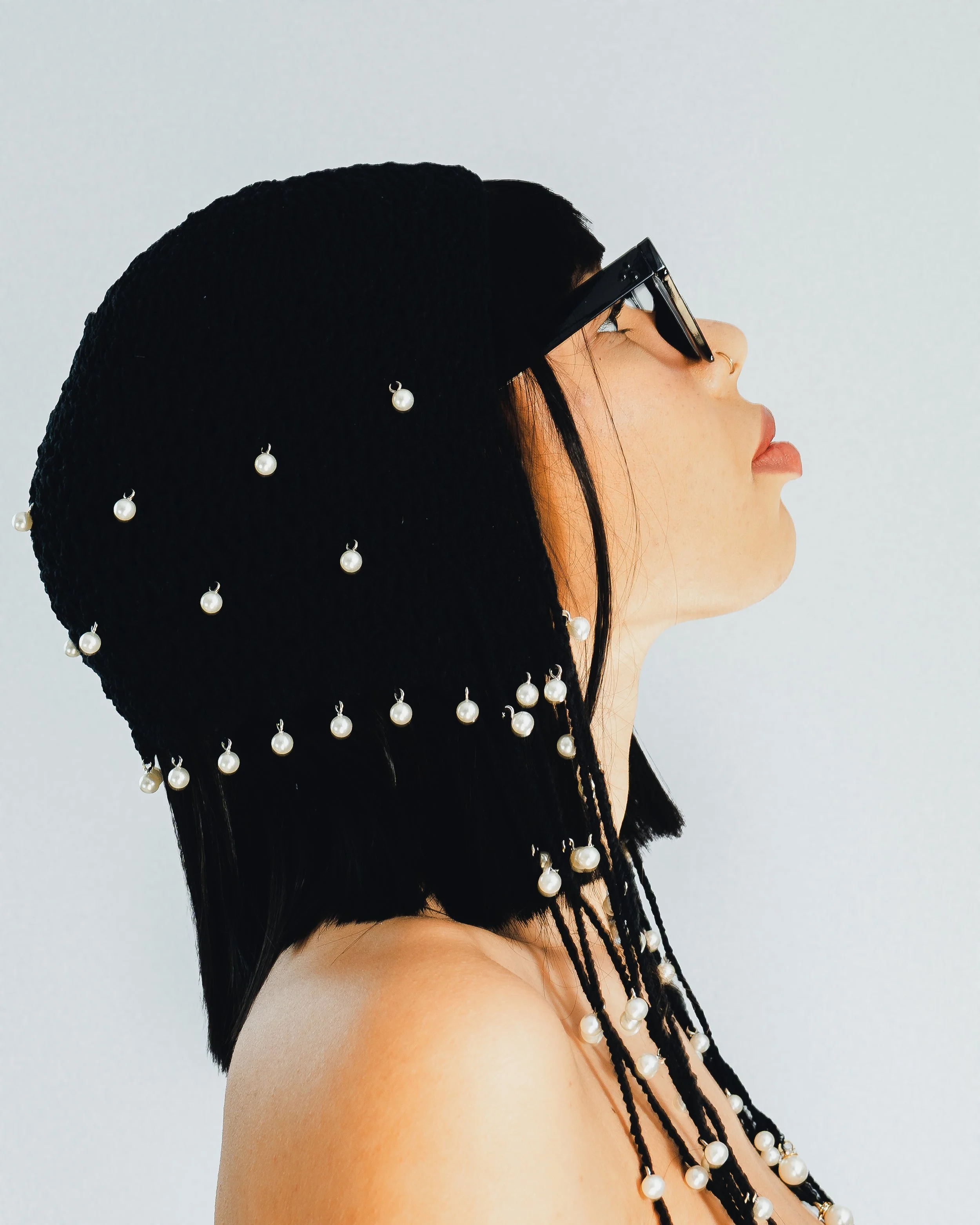 Profil d'une femme avec un sombrero noir orné de perles, portant des lunettes noires et un piercing au nez, contre un fond gris clair.