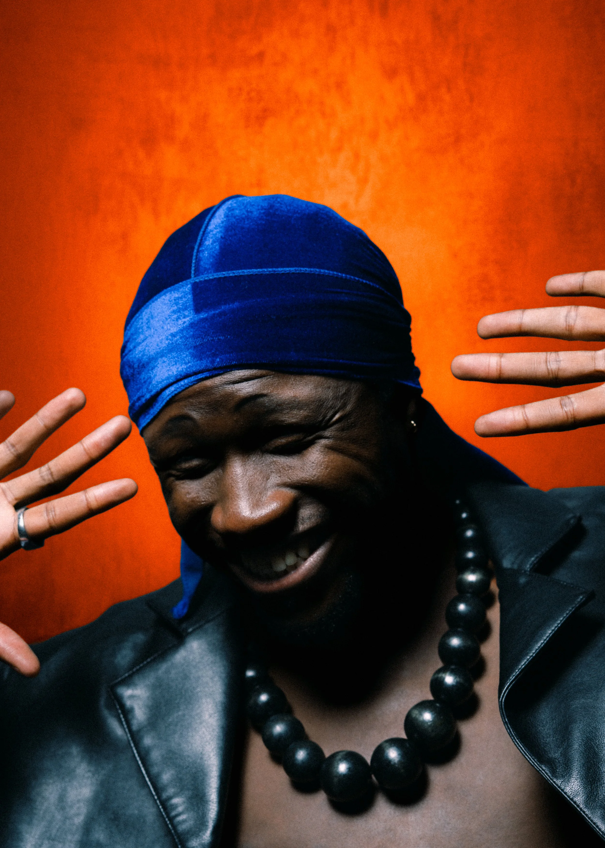 Une femme souriante portant un foulard bleu, un collier de perles noirs et un ensemble en cuir noir, avec un fond orange.
