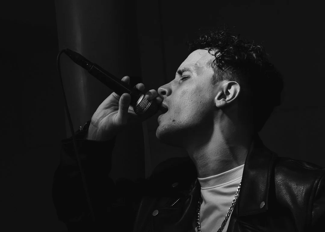 Un jeune homme chantant ou parlant dans un microphone, en noir et blanc, avec une expression concentrée.