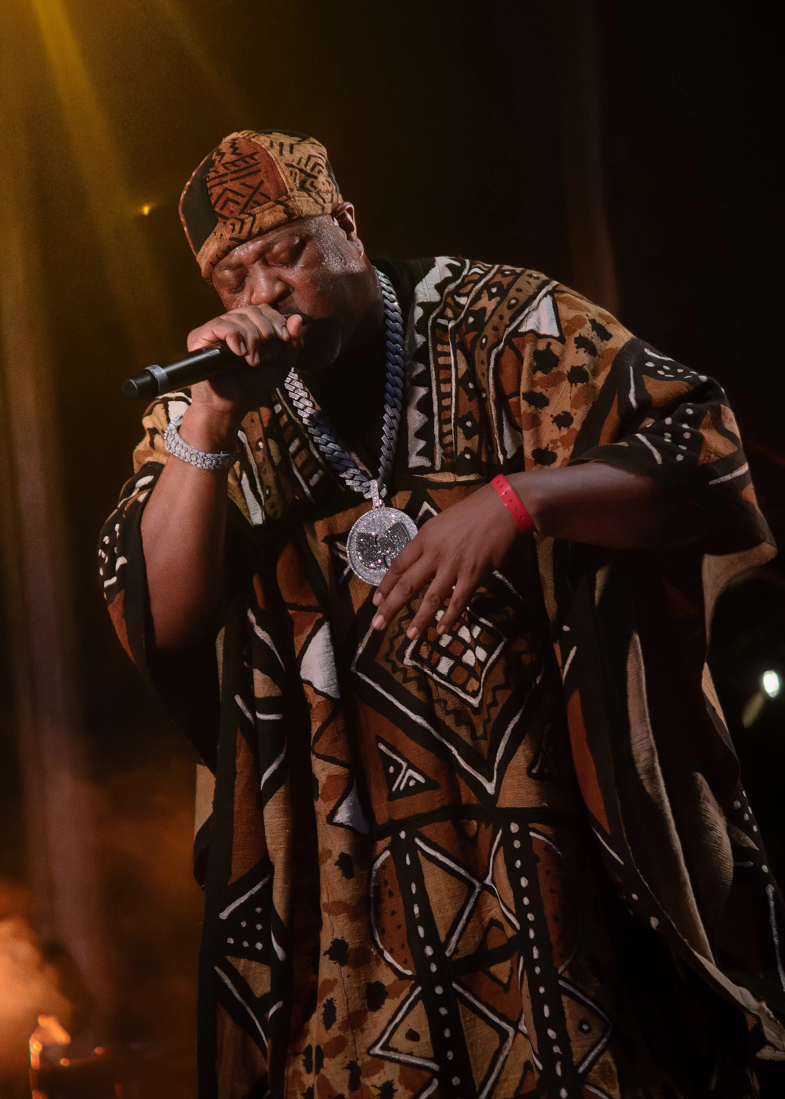Un homme porte un vêtement traditionnel africain et un chapeau avec un motif complexe. Il tient un microphone et porte un collier en chaîne et un bracelet rouge.
