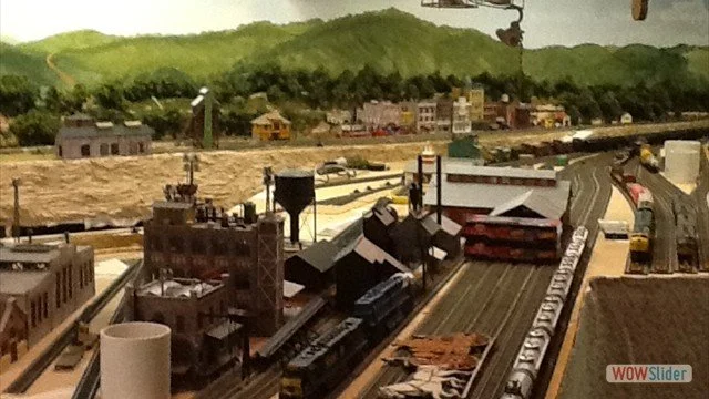 Model train layout with tracks, buildings, and landscape, including a hill with houses in the background.