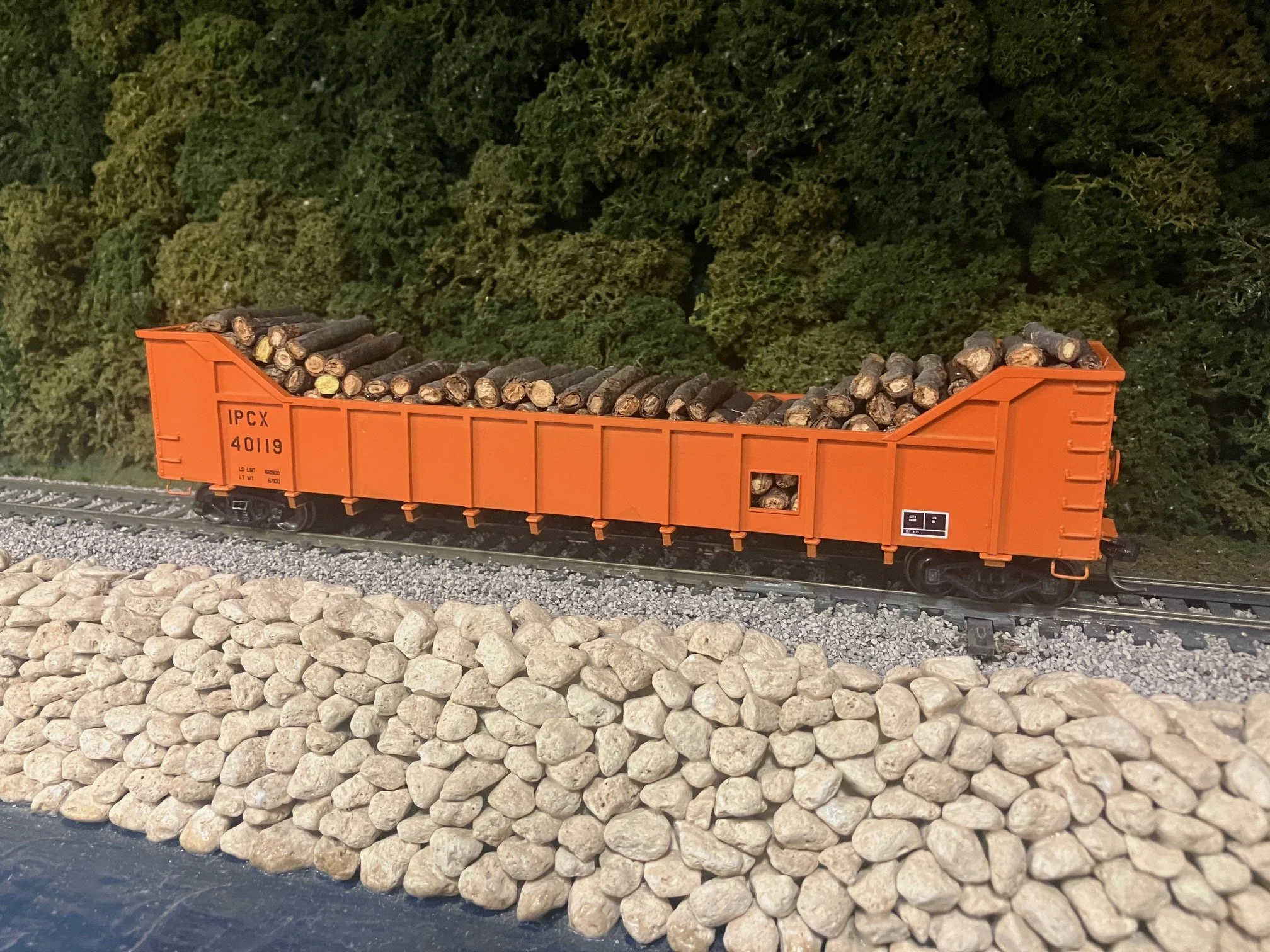 A model train flatcar filled with small stones, on a track with gravel and rocks, against a backdrop of dense green foliage.
